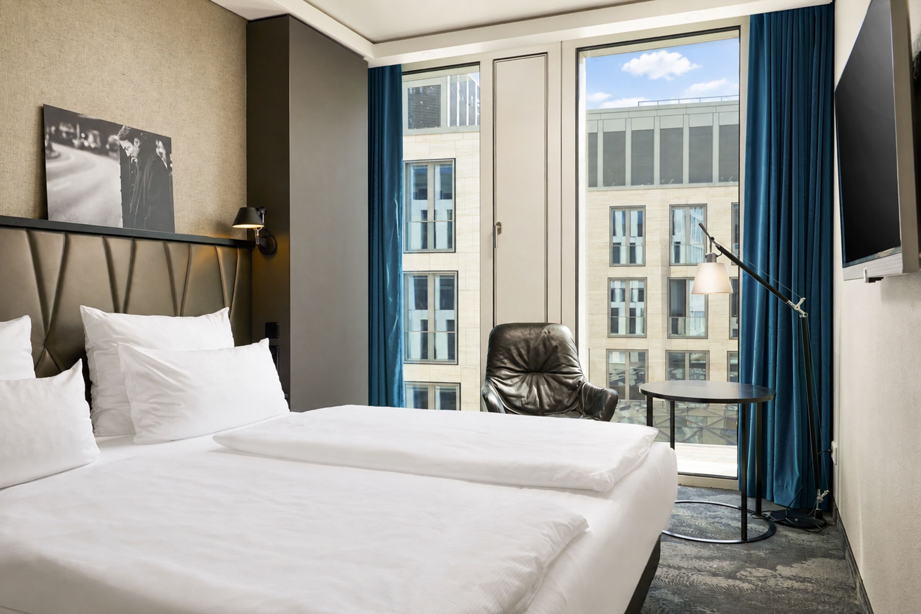 Modern eingerichtetes Hotelzimmer mit großem Doppelbett, bequemen Sessel, Panoramafenstern mit Blick auf umliegende Gebäude und sanfter Beleuchtung für entspannten Aufenthalt.
