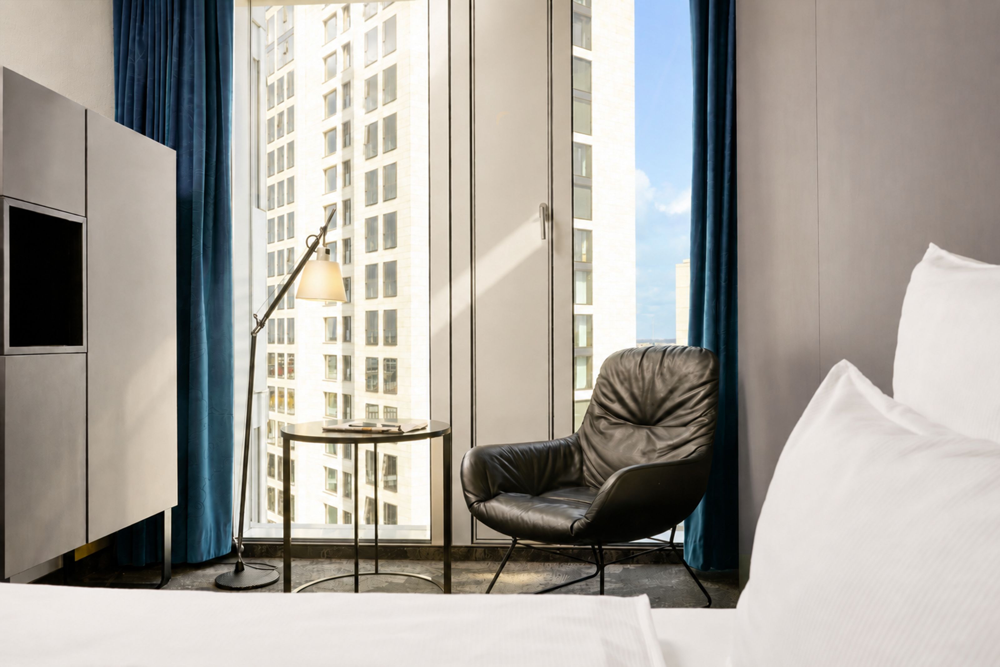 Hell eingerichtetes Hotelzimmer mit großem Fenster, Leder-Sessel, Beistelltisch und Leselampe, Blick auf Hochhäuser.