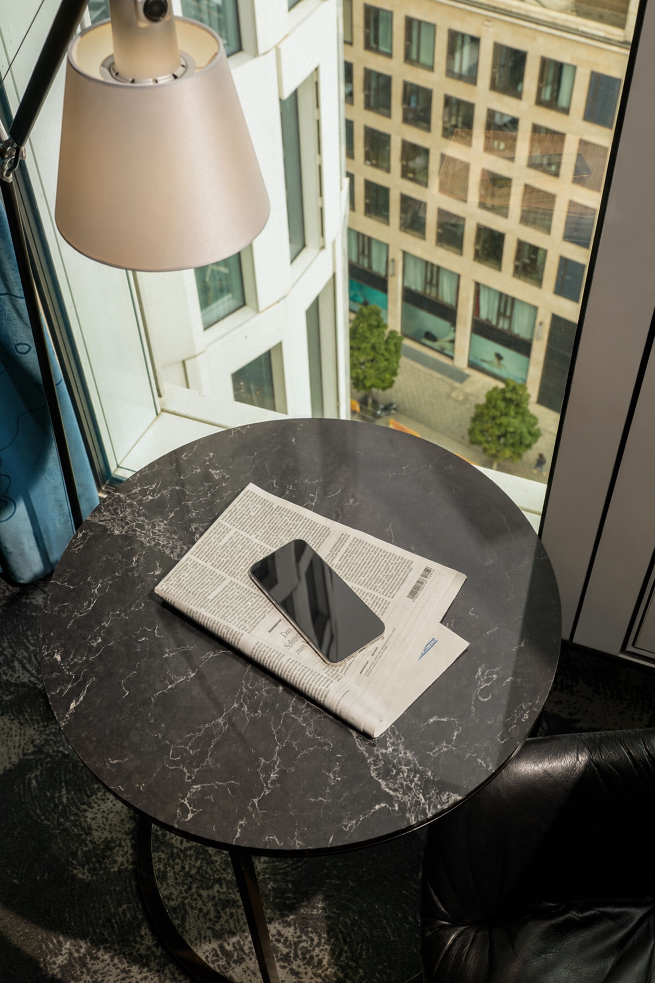 Runder Marmortisch am Fenster mit einer Tageszeitung und einem Smartphone, darüber eine moderne Lampe, Blick auf Bürogebäude und städtischen Innenhof.