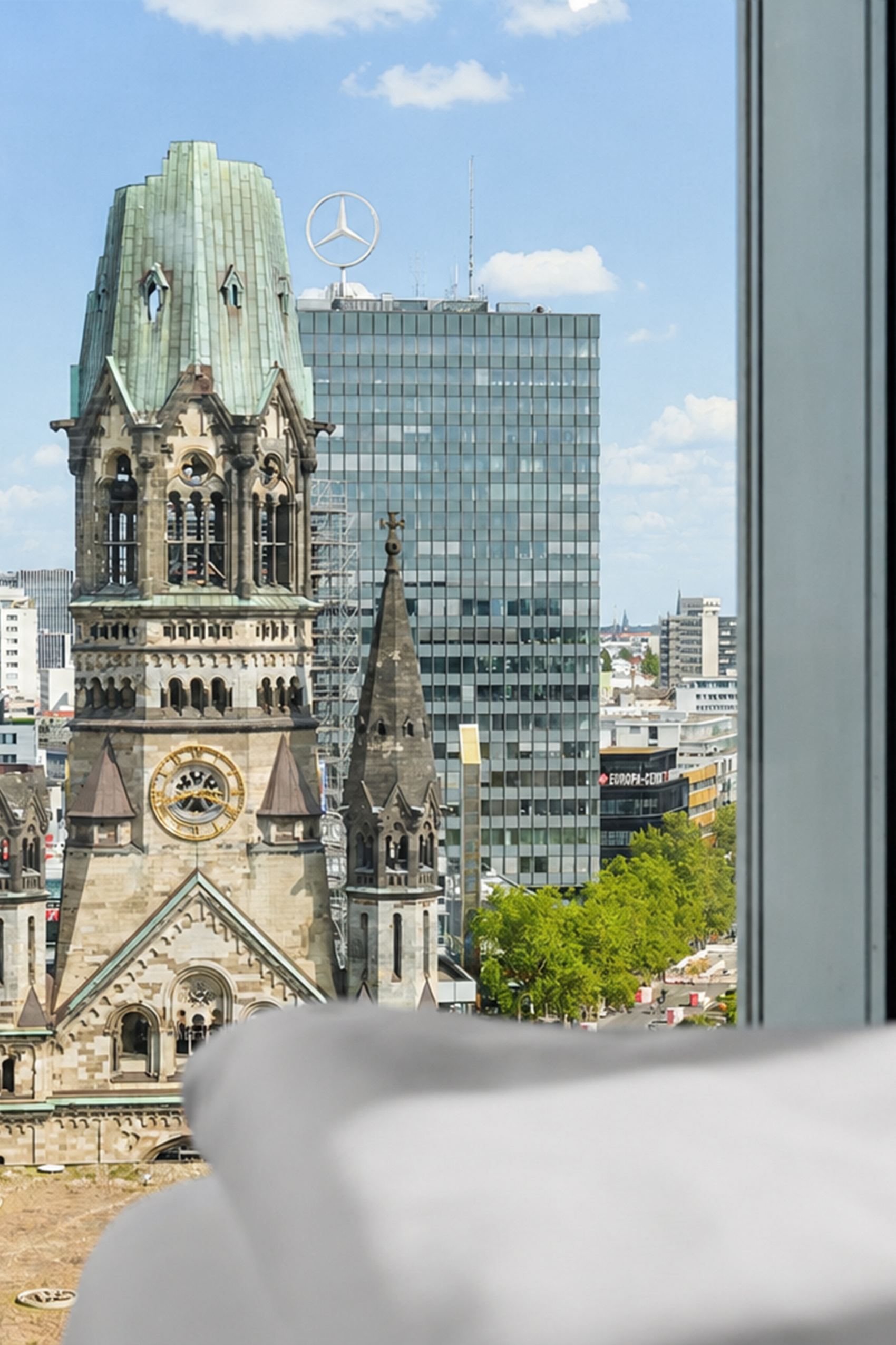 Blick aus einem Hotelzimmerfenster auf die Kaiser-Wilhelm-Gedächtniskirche und moderne Gebäude in der Berliner Innenstadt bei klarem Himmel.