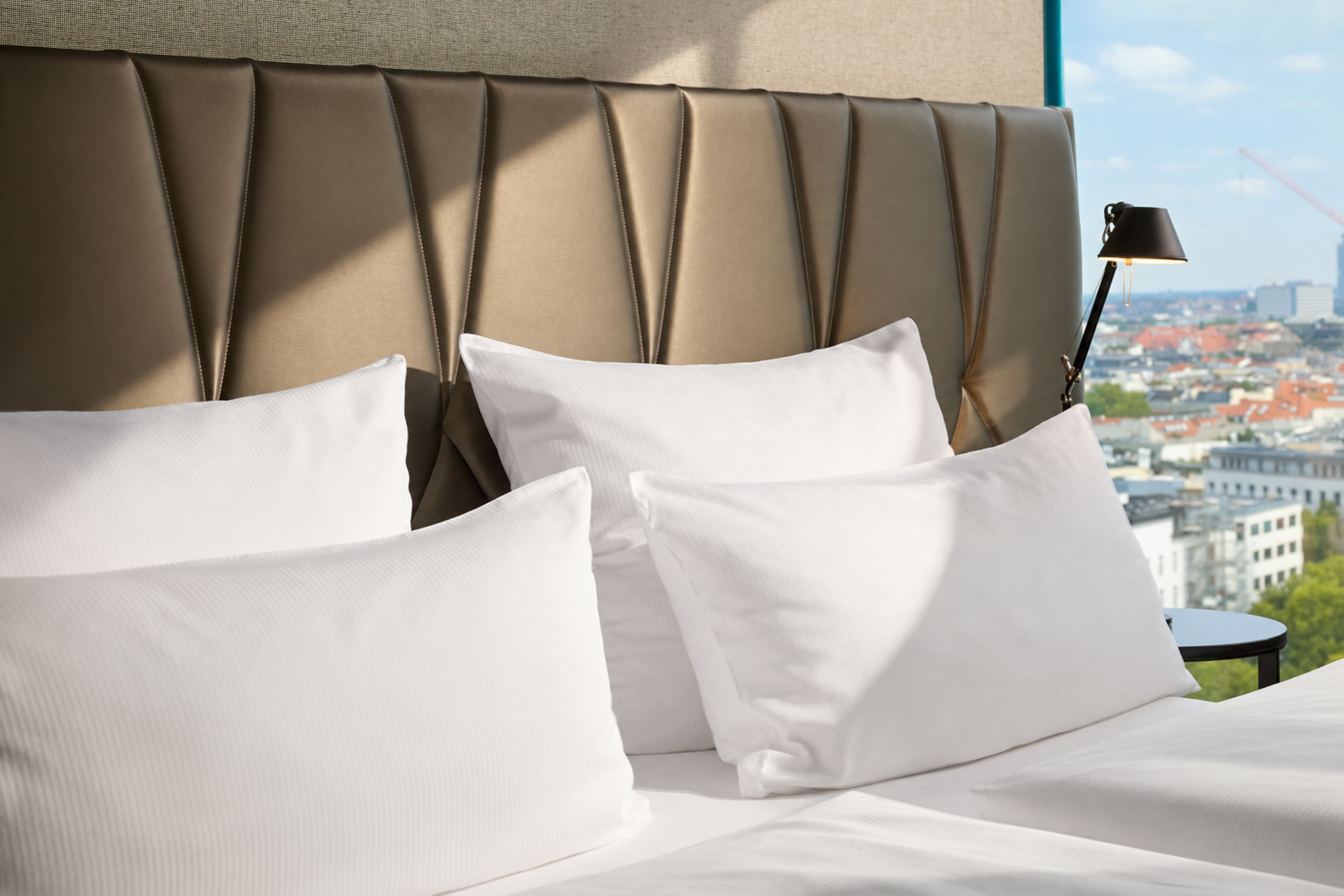 Helles, modern eingerichtetes Hotelzimmer mit großem, gepolstertem Kopfteil, mehreren weißen, flauschigen Kissen und Ausblick auf die Stadt durch ein großes Fenster.