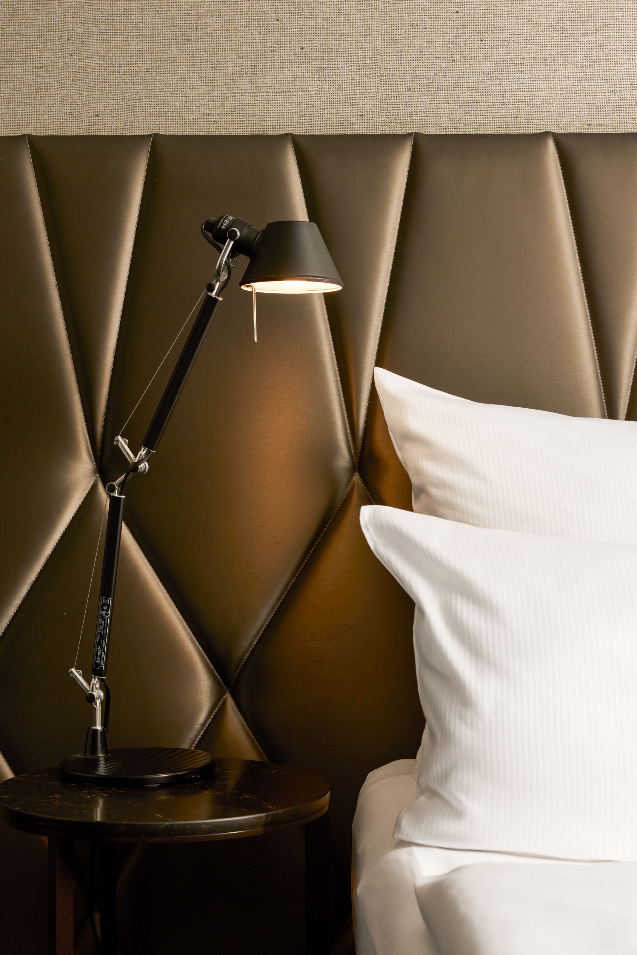 Elegantes Hotelzimmer mit gepolstertem Kopfteil, weißer Bettwäsche und einer modernen, schwarzen Nachttischlampe auf einem runden Nachttisch, die eine gemütliche und ansprechende Atmosphäre für erholsamen Schlaf schafft.