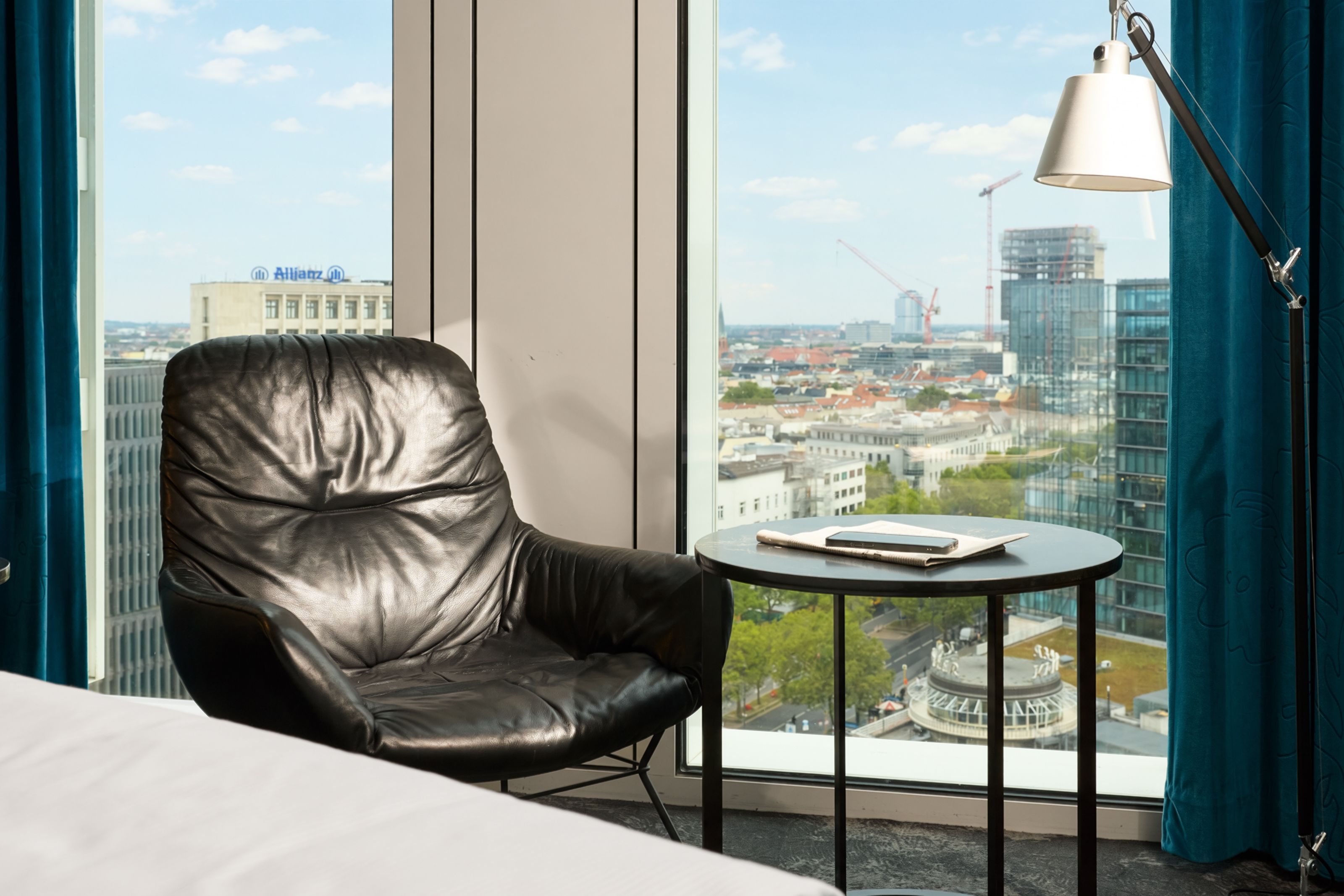 Blick aus einem Hotelzimmer mit großem Fenster auf die Stadt, ein bequemer schwarzer Ledersessel und ein kleiner runder Tisch mit Notizbuch und Stift laden zum Entspannen und Arbeiten ein.