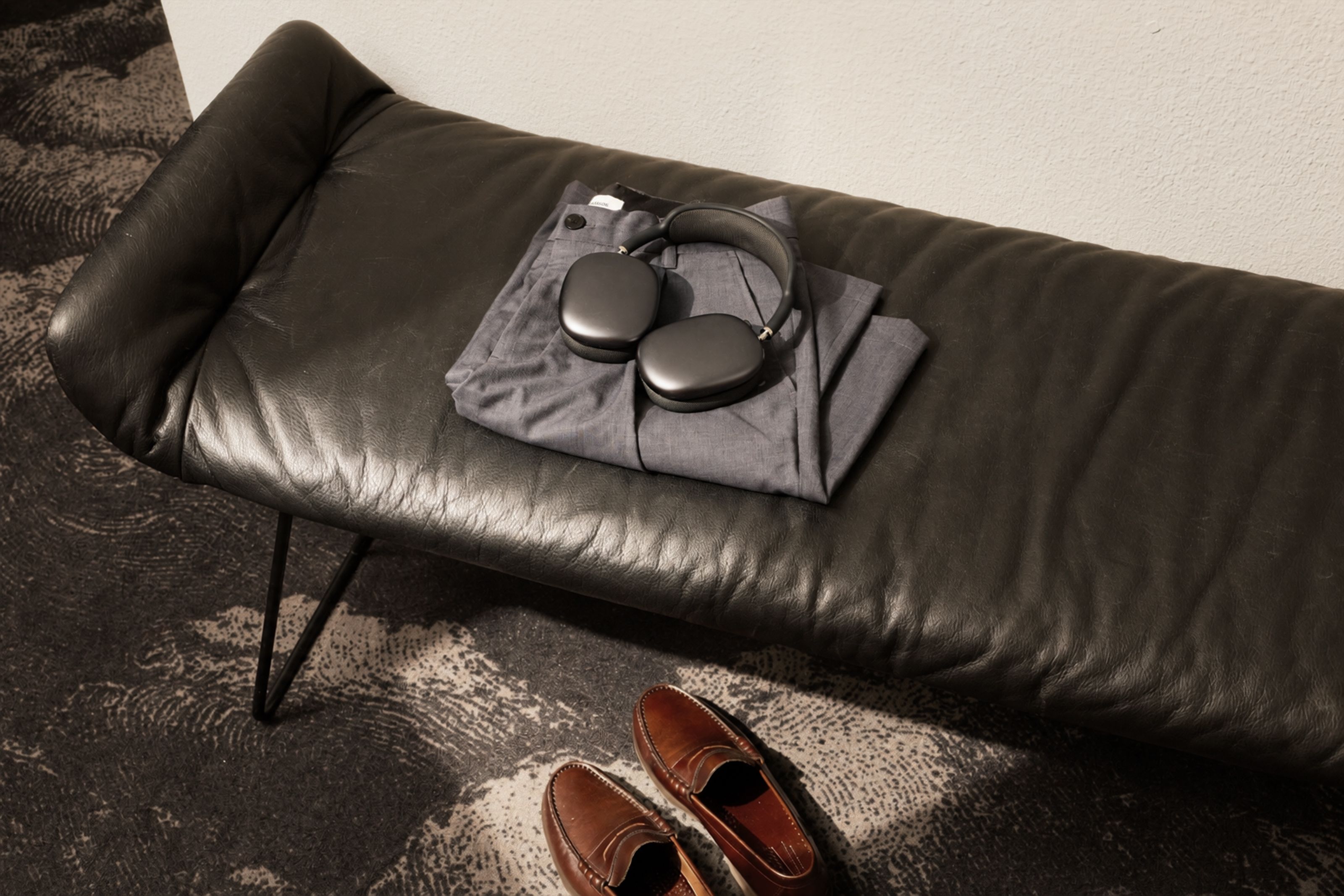 Bequeme schwarze Ledercouch mit ordentlich gefalteter Kleidung und drahtlosen Kopfhörern darauf, daneben elegante braune Herrenlederchuhe auf gemustertem Teppich.