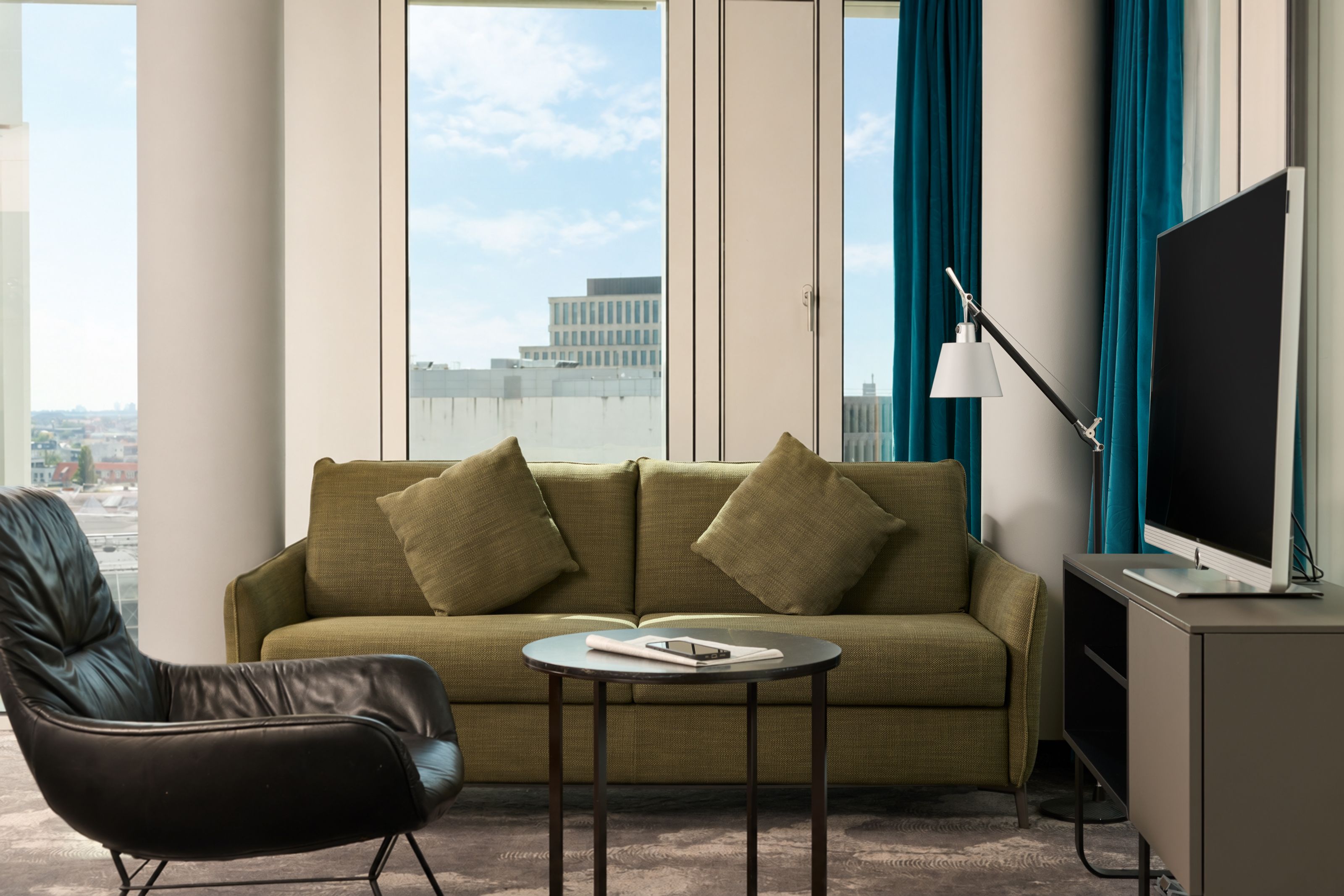 Moderner Wohnbereich mit grünem Sofa, zwei Kissen, kleinem runden Tisch und gemütlichem schwarzem Sessel vor großen Fenstern mit Stadtblick, TV und dekorativem Leselicht.