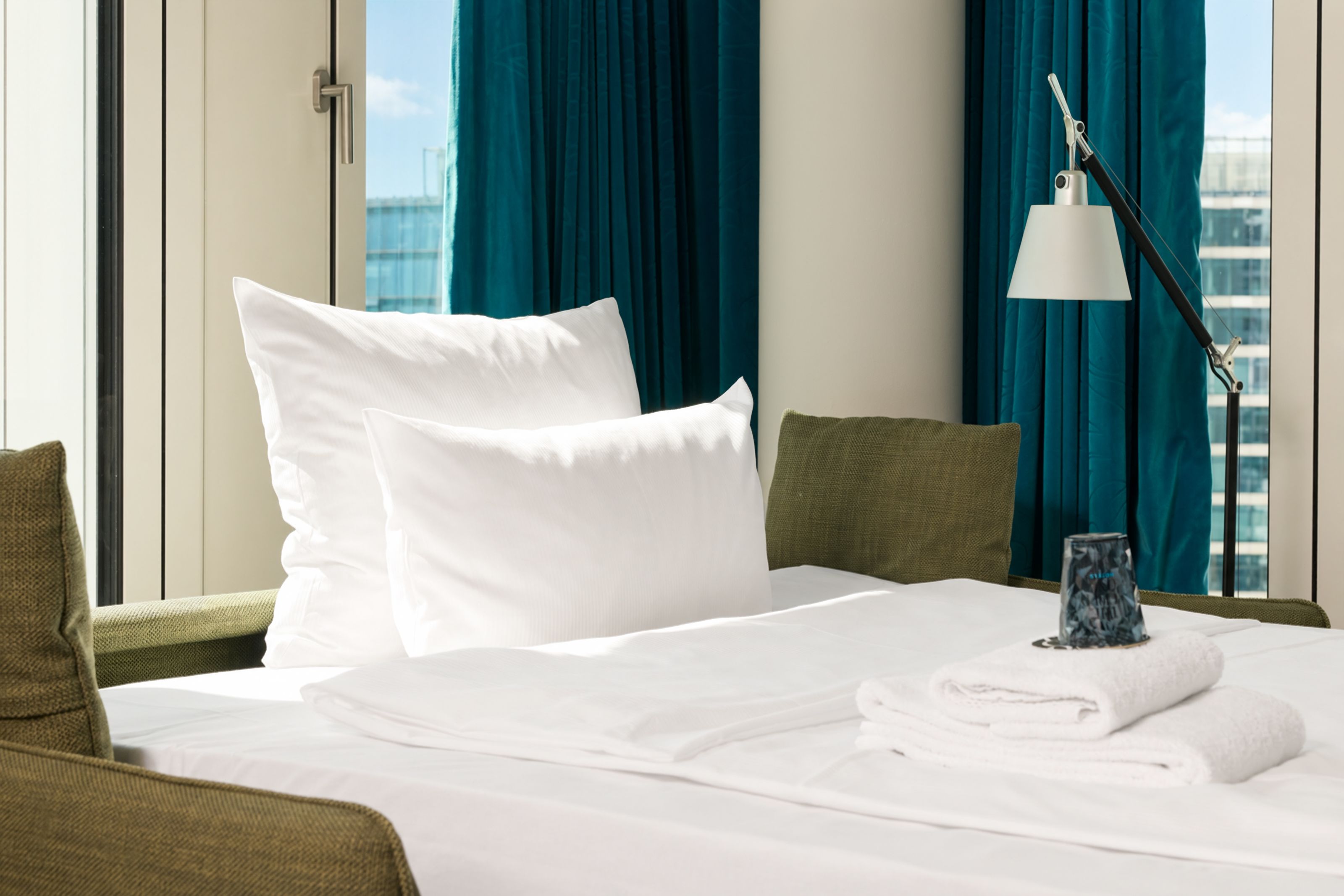 Modern eingerichtetes Hotelzimmer mit großem, weiß bezogenem Bett, frischen Handtüchern und Decken, grünen Sesseln und großen Fenstern mit Blick auf die Stadt. Helle, ruhige Atmosphäre ideal für Erholung und komfortables Wohnen.
