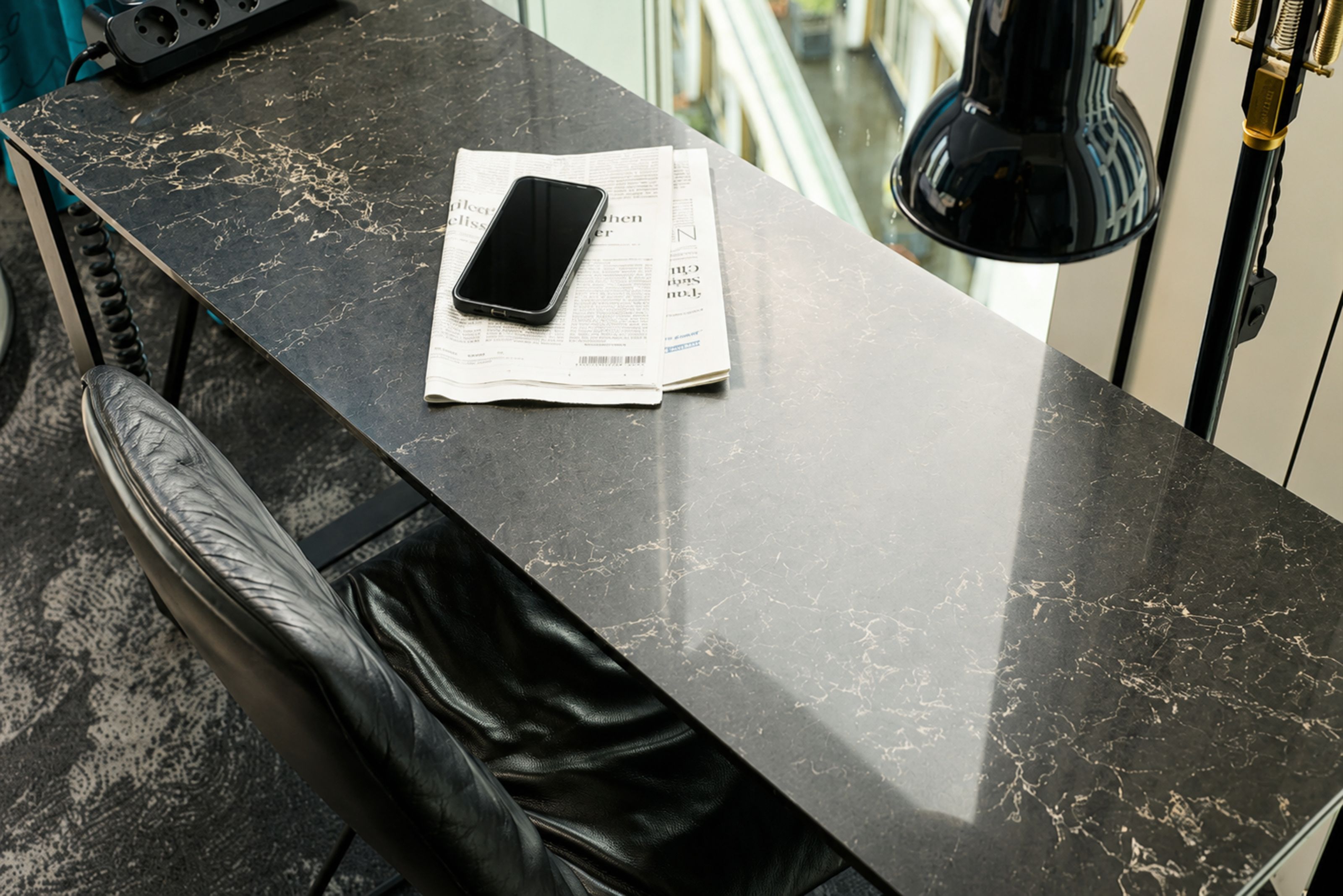 Schwarzer Marmor-Schreibtisch mit Zeitung und Smartphone, schwarzer Lederstuhl und Stehlampe, neben Fenster mit Blick auf Stadt. Ambiente für konzentriertes, stilvolles Arbeiten im Hotelzimmer.