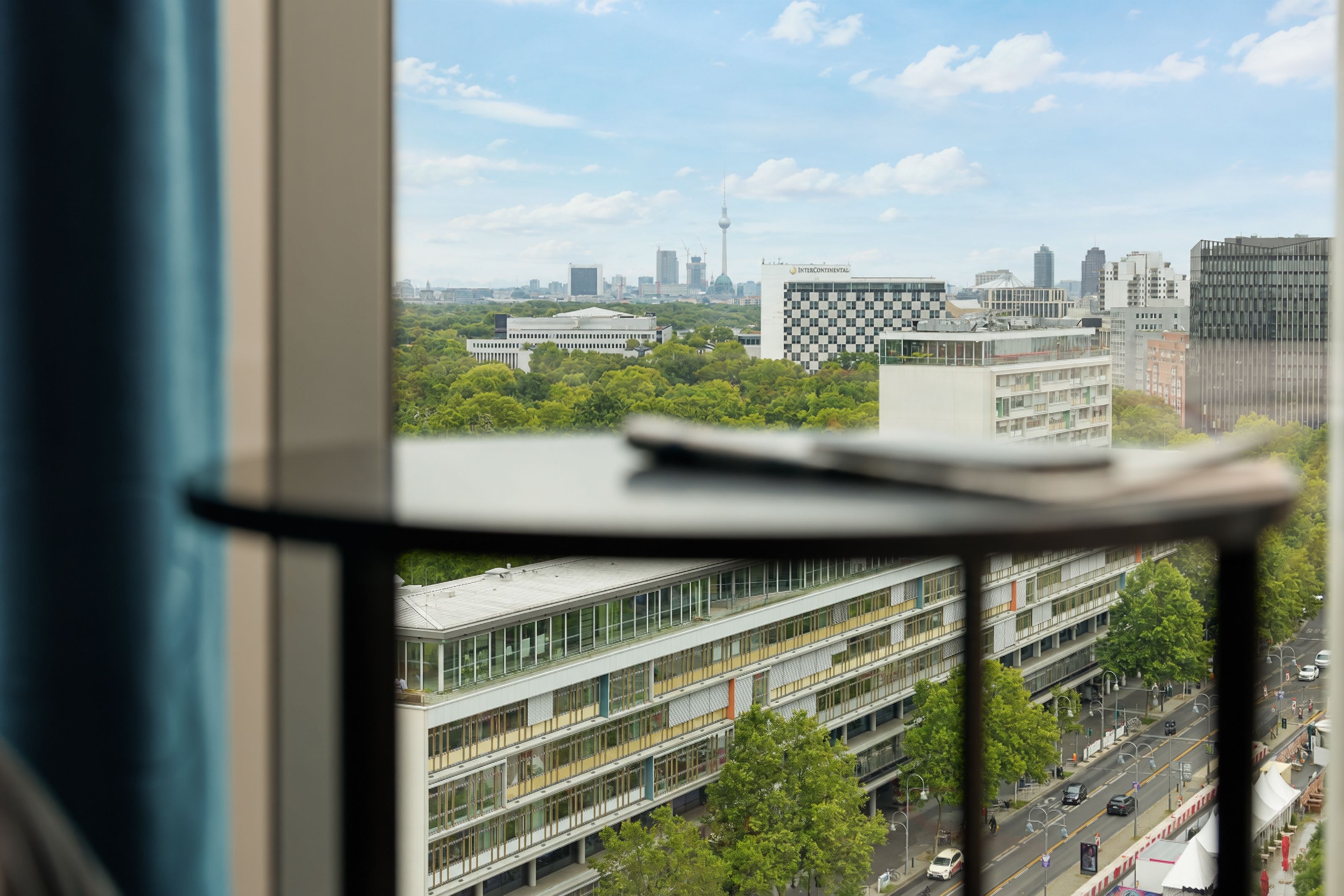 Blick aus einem Hotelfenster auf Berlins Stadtzentrum mit grünen Parkflächen, modernen Gebäuden und dem Fernsehturm am Horizont bei klarem Himmel.