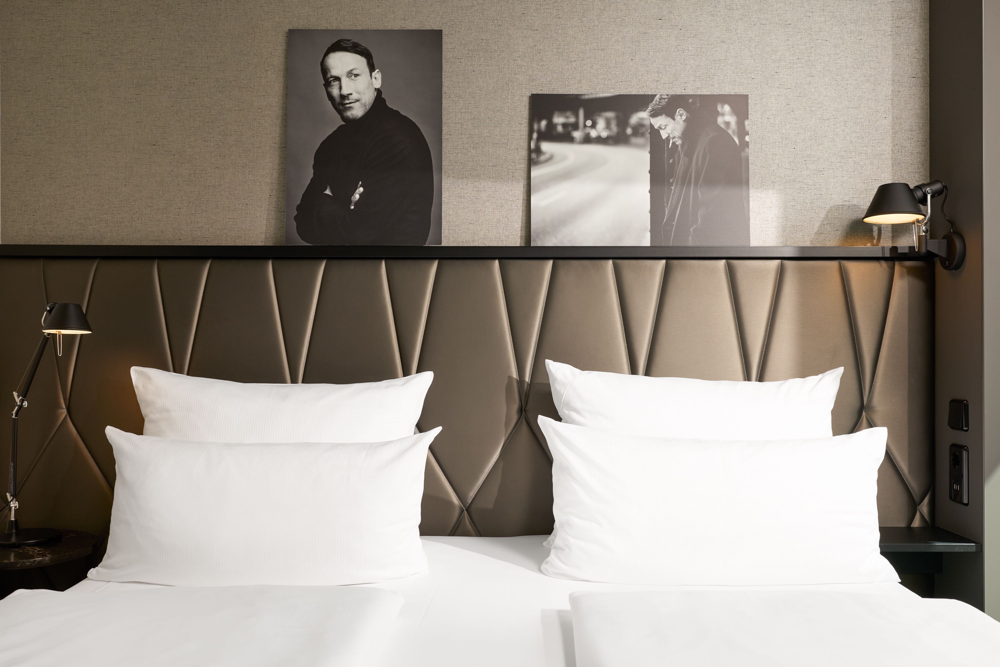 Modern eingerichtetes Hotelzimmer mit Doppelbett, weißer Bettwäsche, gepolstertem Kopfteil und schwarz-weißen Fotografien an der Wand für eine stilvolle Atmosphäre.