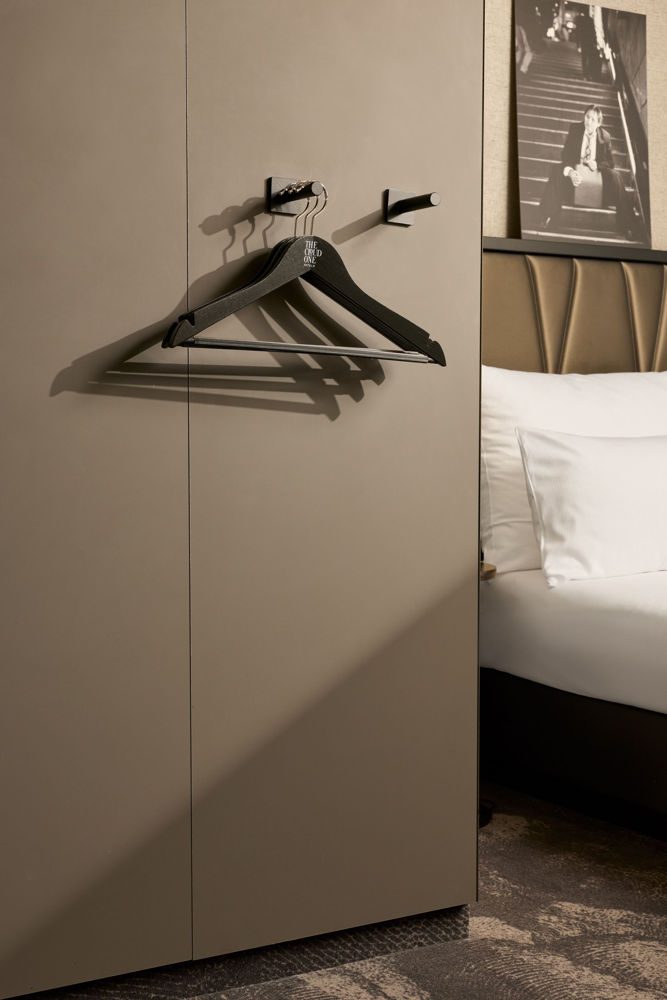 Schlichte Garderobe mit schwarzen Kleiderbügeln an der Wand, neben einem komfortablen Bett mit weißer Bettwäsche, in einem elegant und modern eingerichteten Hotelzimmer.