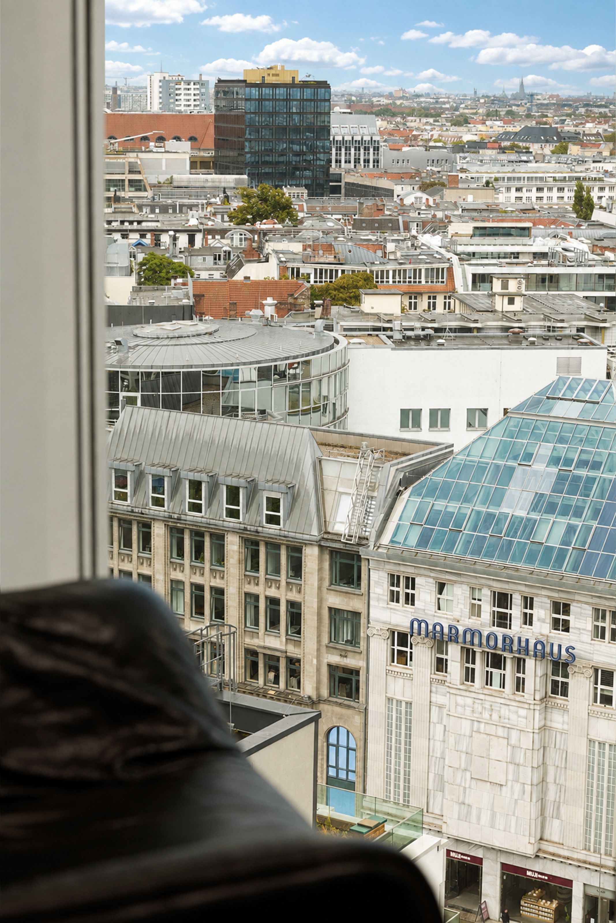 Blick aus einem Hotelfenster auf die Dächer und Fassaden moderner und historischer Gebäude der Stadt, darunter das Marmorshaus mit gläsernem Dach, bei klarem Himmel.