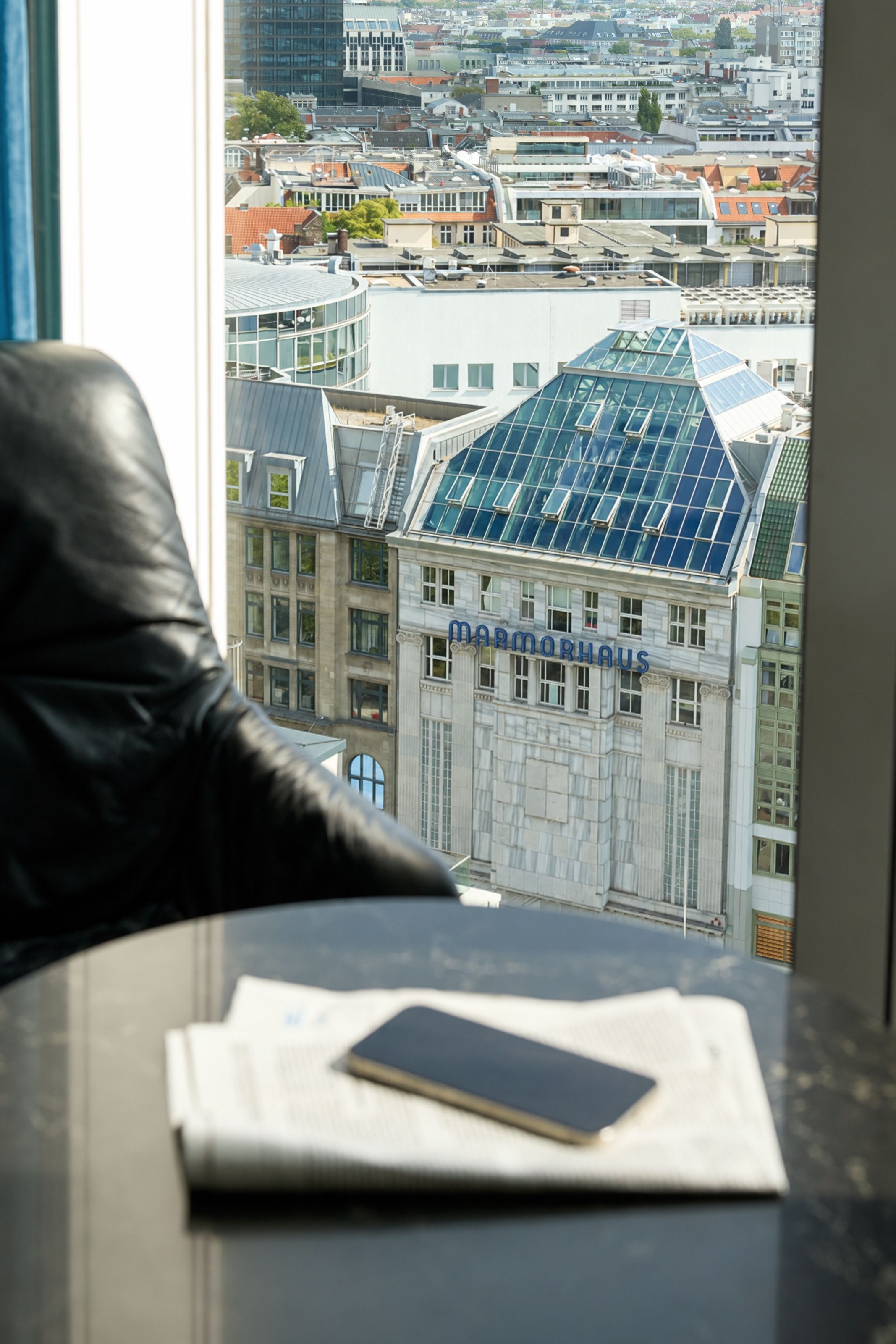 Blick aus einem Hotelzimmerfenster auf das Markthaus-Gebäude mit Glasdach und die Stadt dahinter, im Vordergrund ein Tisch mit Zeitungen und einem Smartphone.