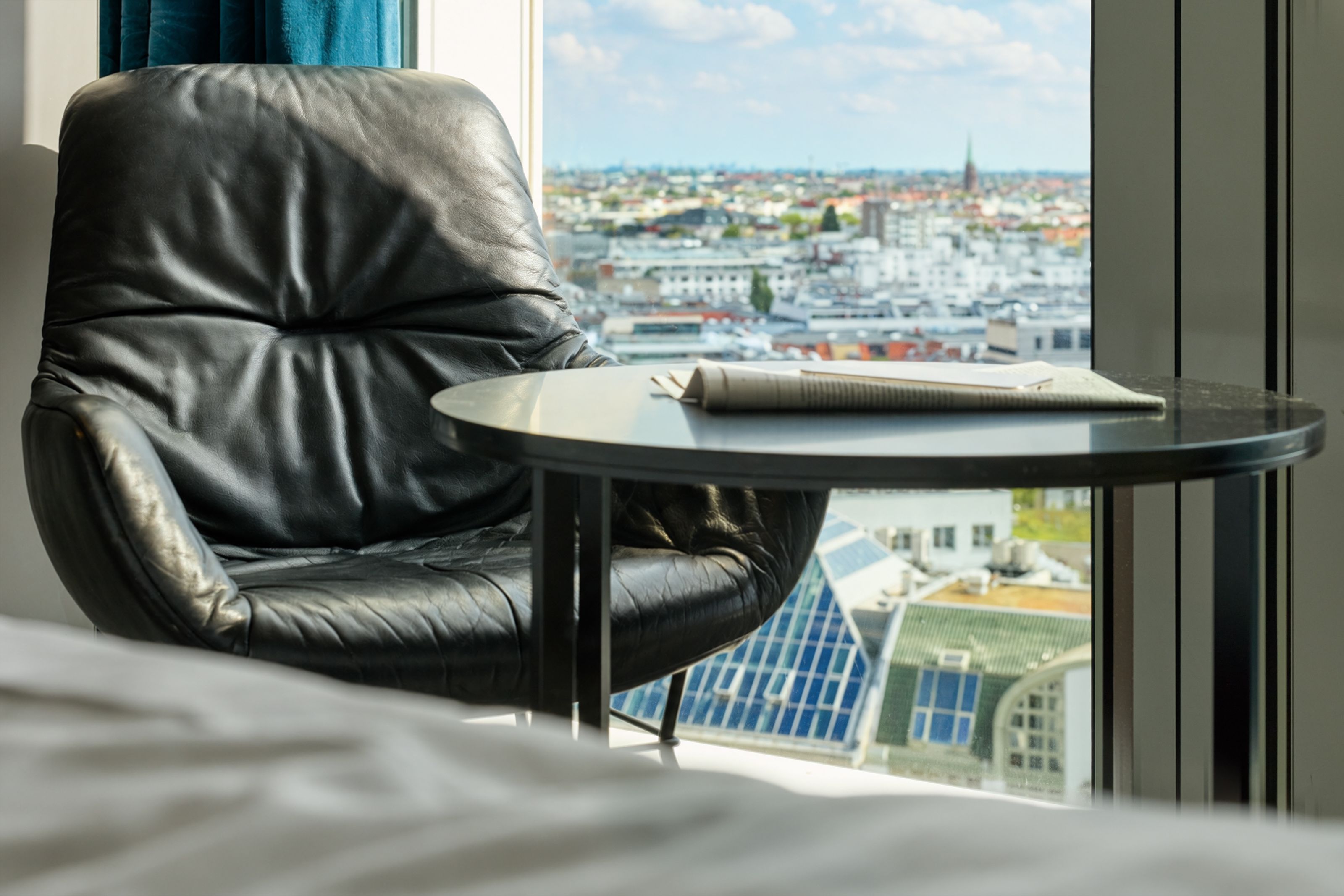 Bequemer Ledersessel und runder Tisch vor großem Fenster mit Blick über eine Stadtlandschaft, helle Tageslichtstimmung vermittelt Komfort und Entspannung im urbanen Hotelzimmer.