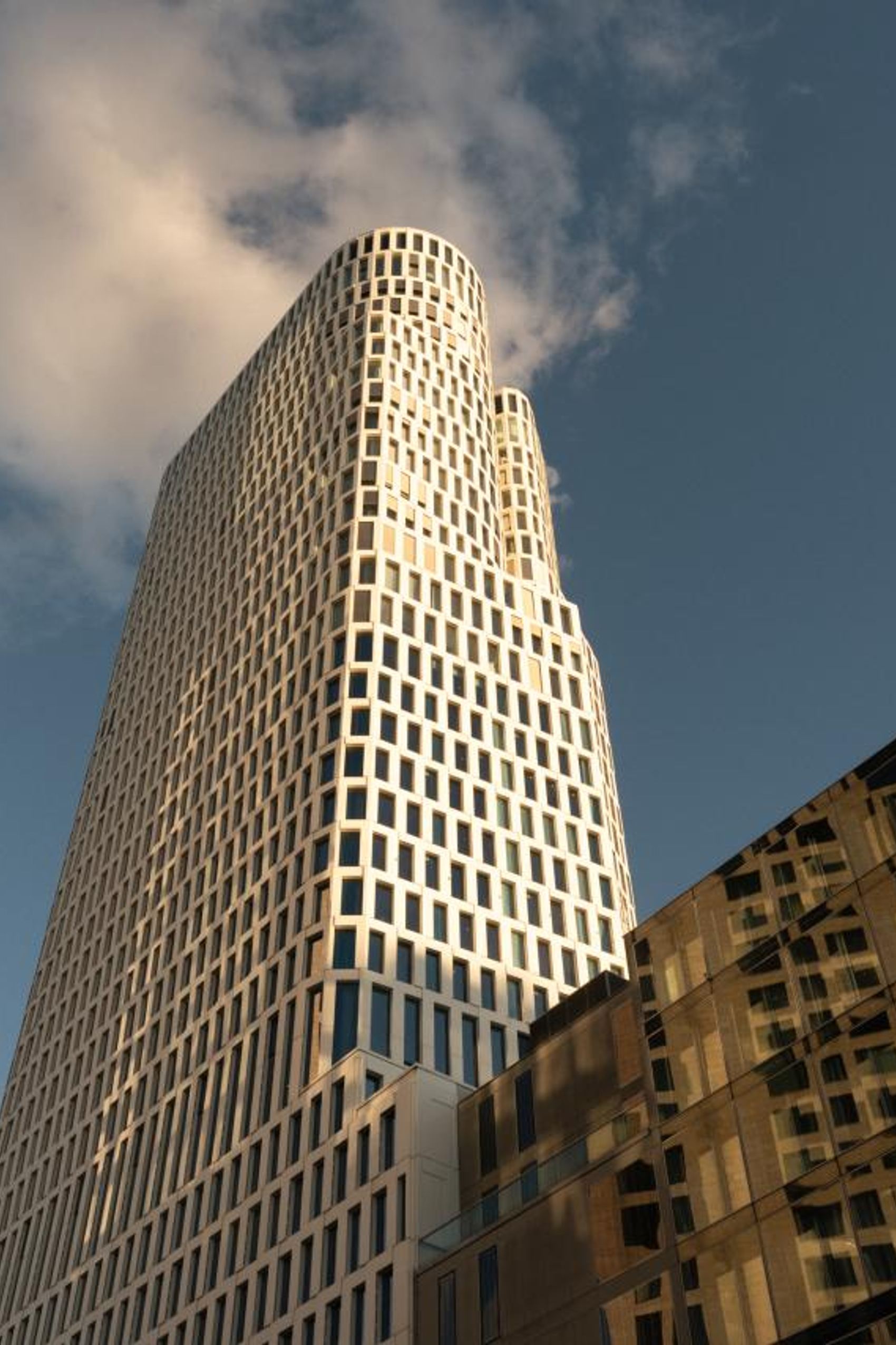 Moderner Wolkenkratzer mit wellenförmiger Fassade, aufgenommen bei warmem Tageslicht, vor blauem Himmel mit einigen Wolken, umgeben von weiteren Gebäuden. Ideal für Geschäftsreisende, die urbanes Ambiente und zeitgemäße Architektur schätzen.
