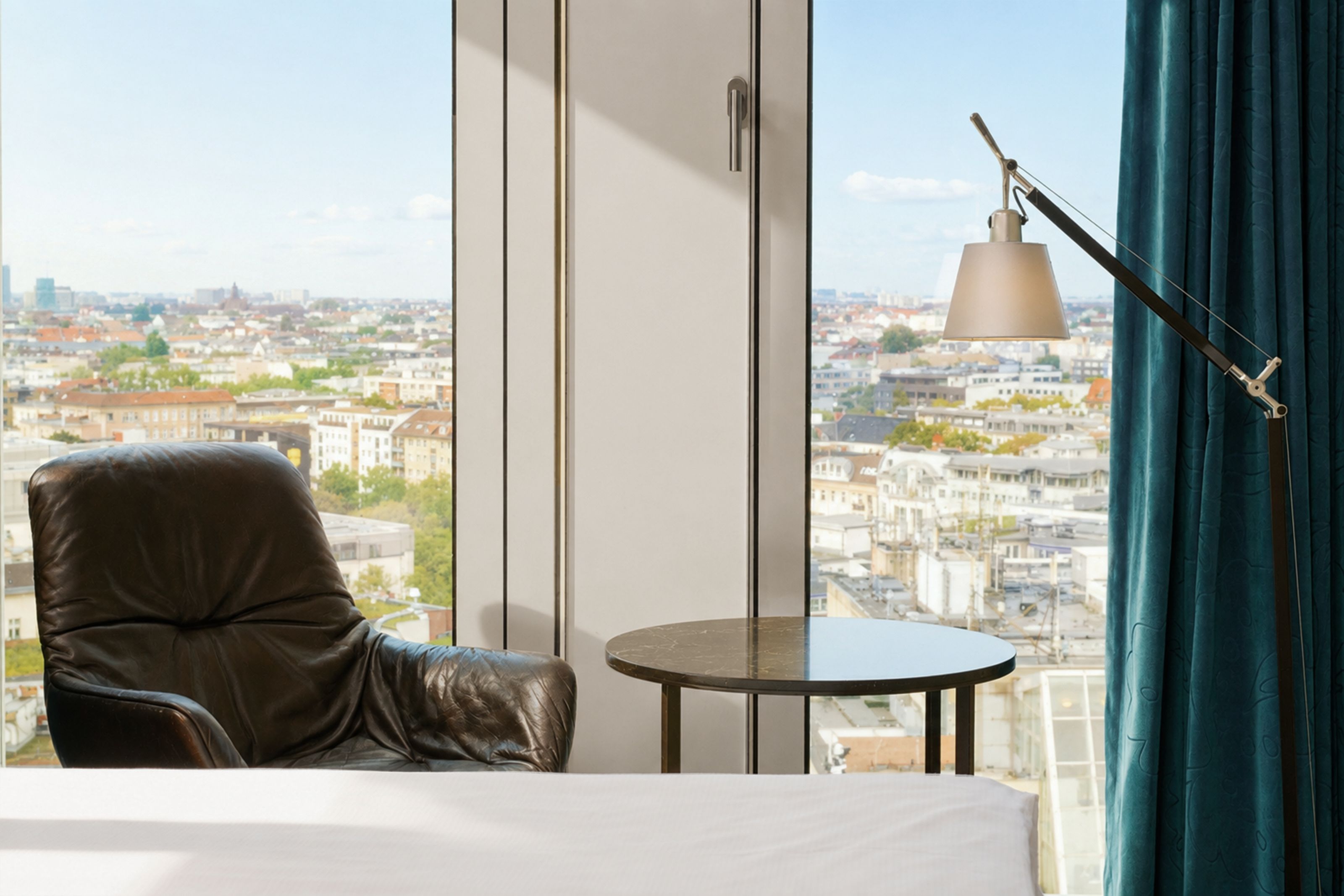 The Cloud One Berlin-Upper West Gemütlicher Sitzbereich mit Lederstuhl, Beistelltisch und Leselampe vor bodentiefen Fenstern, die eine weite Stadtansicht bieten.