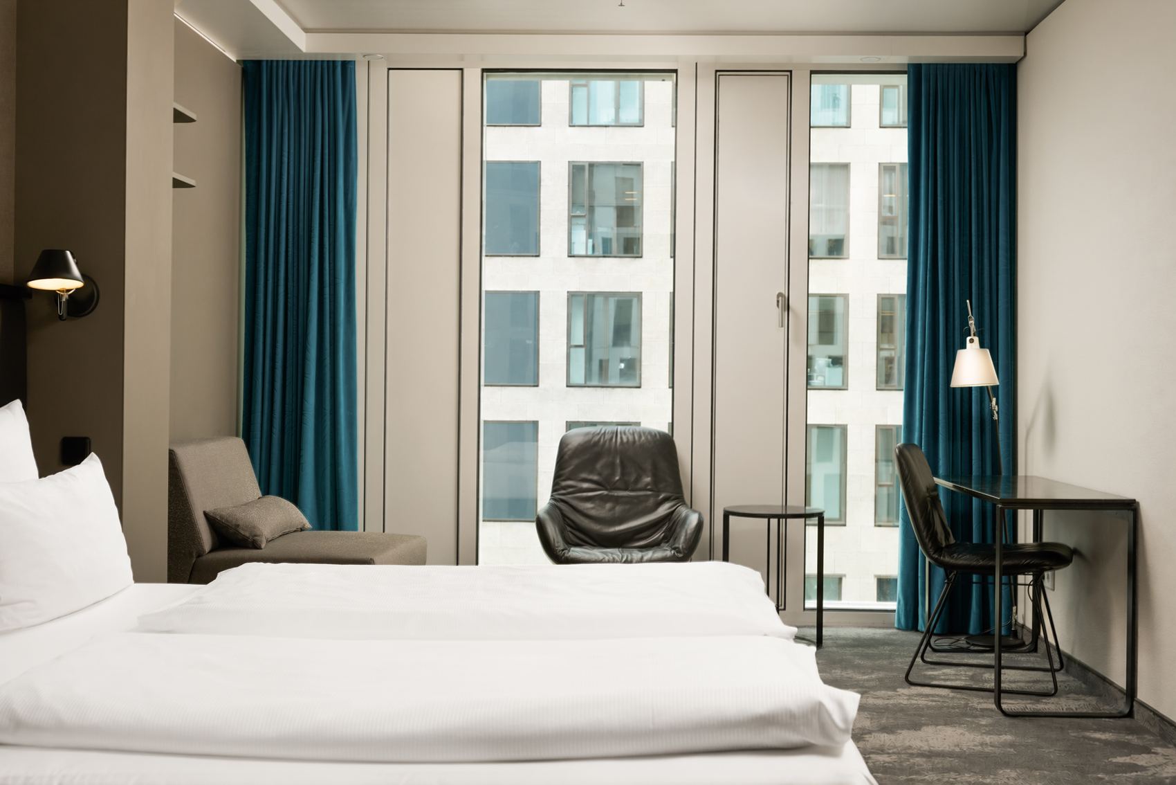 The Cloud One Berlin-Upper West Helles Hotelzimmer mit zwei weißen Betten, einem bequemen Sessel vor großen Fenstern und einem kleinen Arbeitsplatz mit Stuhl, ideal für entspannte oder produktive Momente während des Aufenthalts.