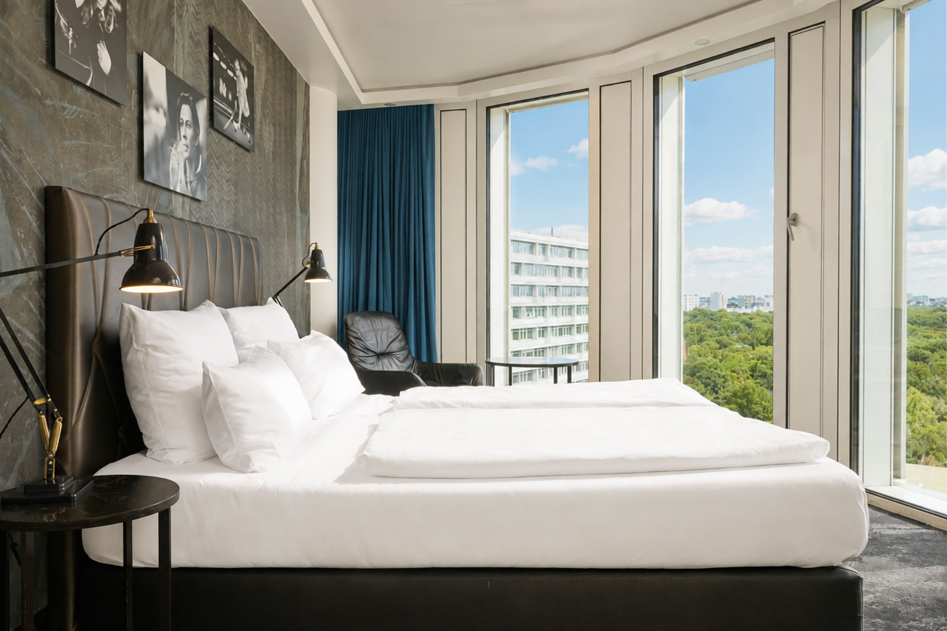 The Cloud One Berlin-Upper West Modern eingerichtetes Hotelzimmer mit großem, komfortablem Bett, Panoramafenstern und Blick auf eine grüne Parklandschaft sowie städtische Gebäude. Helle, klare Atmosphäre mit dunklen Akzenten und einem bequemen Sessel am Fenster.