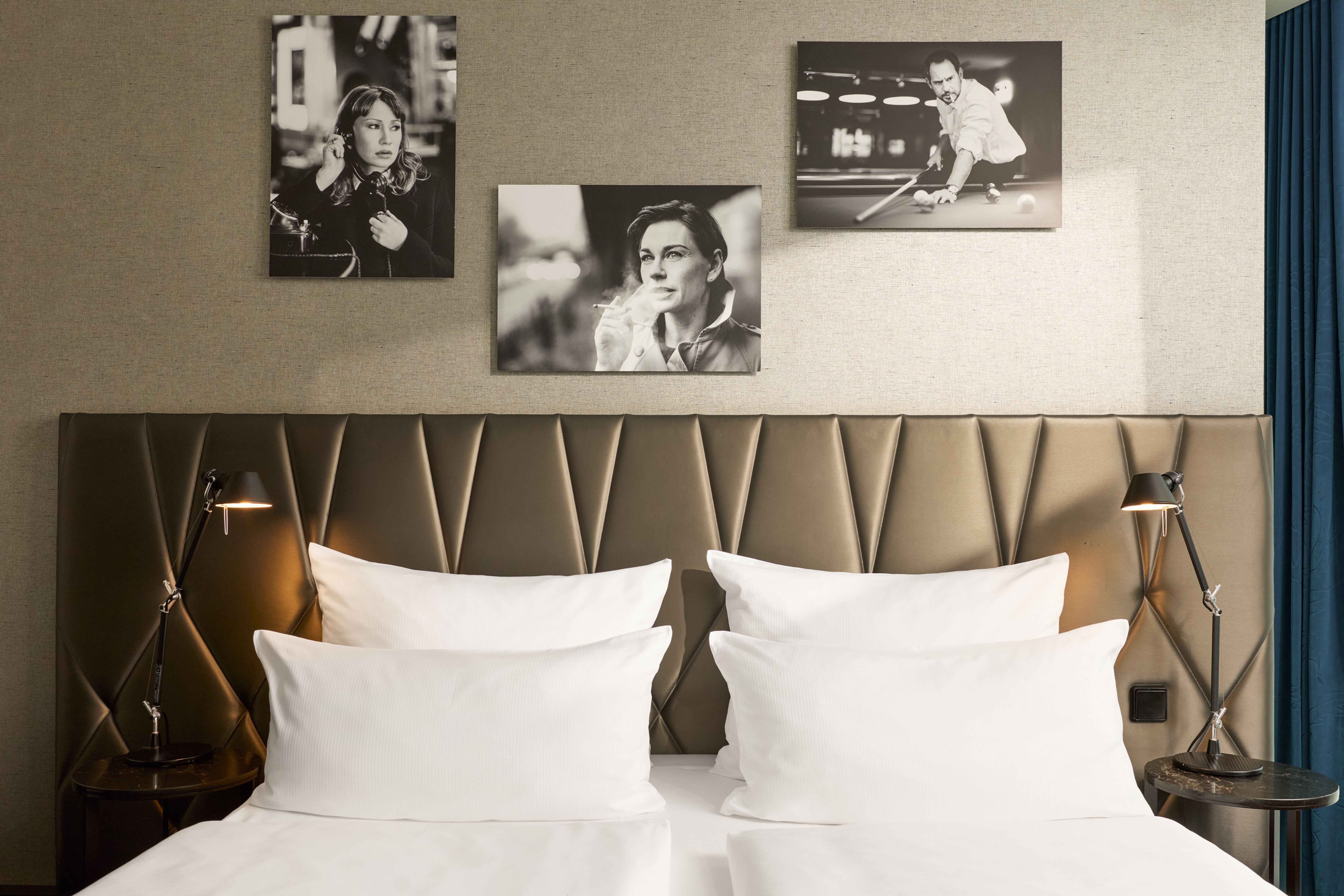 The Cloud One Berlin-Upper West Bett mit gepolstertem Kopfteil, zwei Nachttischlampen und vier weißen Kissen. Oben drei Schwarz-Weiß-Fotografien an der Wand, die entspannte und nachdenkliche Personen zeigen. Moderne, gemütliche Atmosphäre eines Hotelzimmers. The Cloud One Berlin-Upper West Bett mit gepolstertem Kopfteil, zwei Nachttischlampen und vier weißen Kissen. Oben drei Schwarz-Weiß-Fotografien an der Wand, die entspannte und nachdenkliche Personen zeigen. Moderne, gemütliche Atmosphäre eines Hotelzimmers.