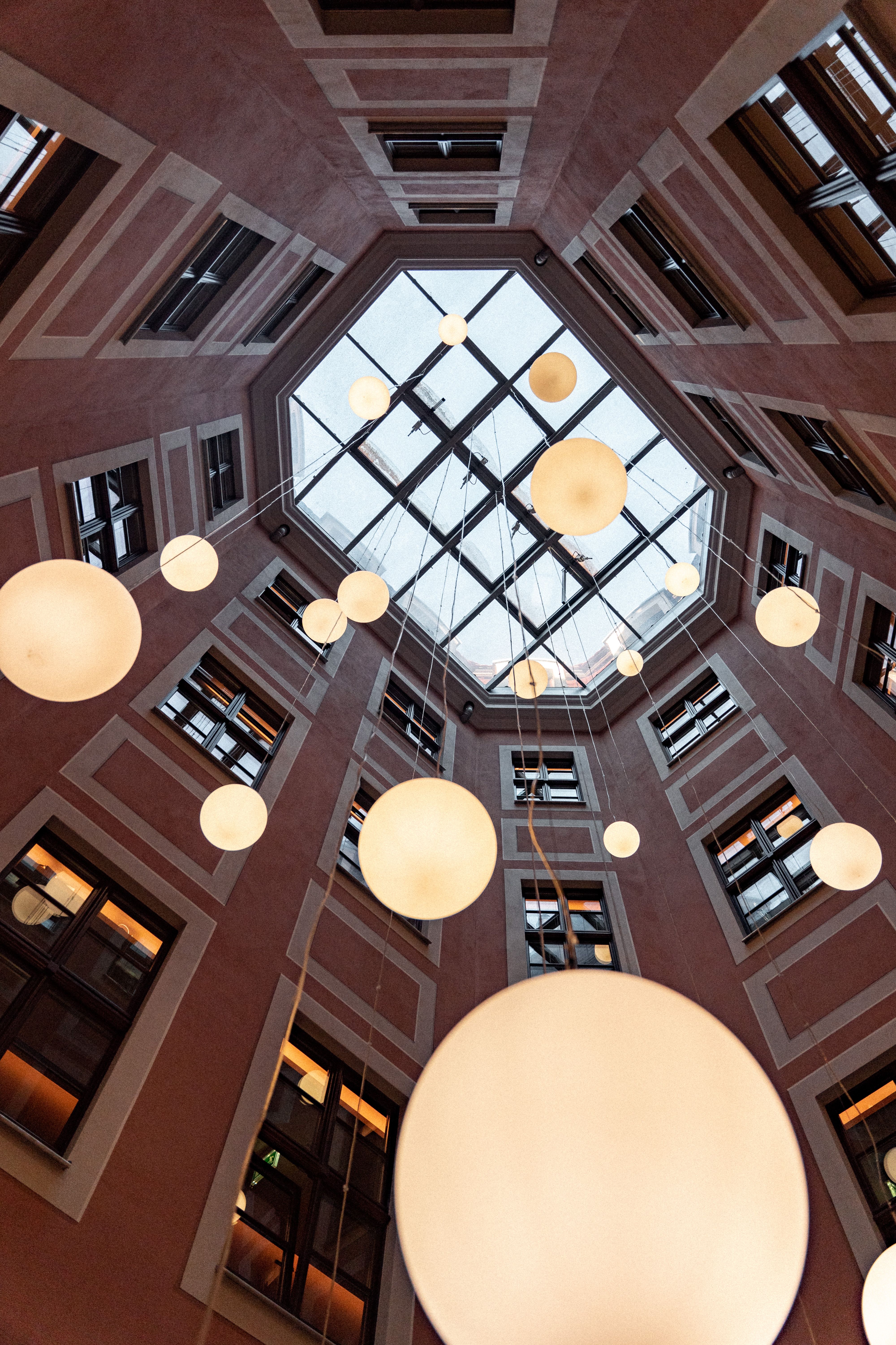 Blick nach oben in einen Innenhof mit rechteckiger Glasdecke, von der mehrere große, runde, warm beleuchtete Kugellampen in verschiedenen Größen hängen. Die umgebenden Wände sind mit Fenstern und einem warmen Farbton gestaltet.