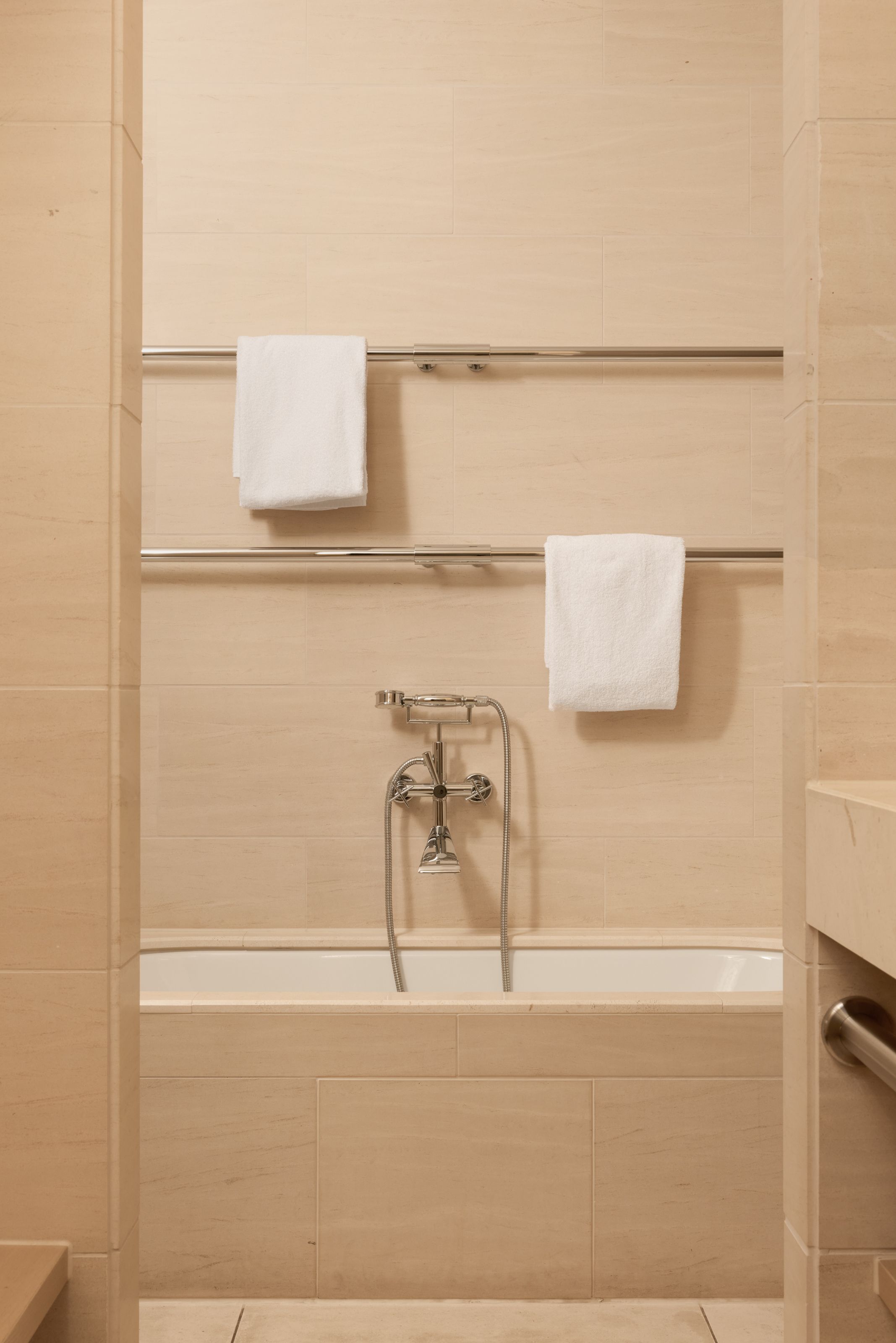 Helle Badewanne mit moderner Wandarmatur und zwei weißen Handtüchern, die an silbernen Handtuchhaltern hängen, in einem beige eingerichteten Badezimmer.