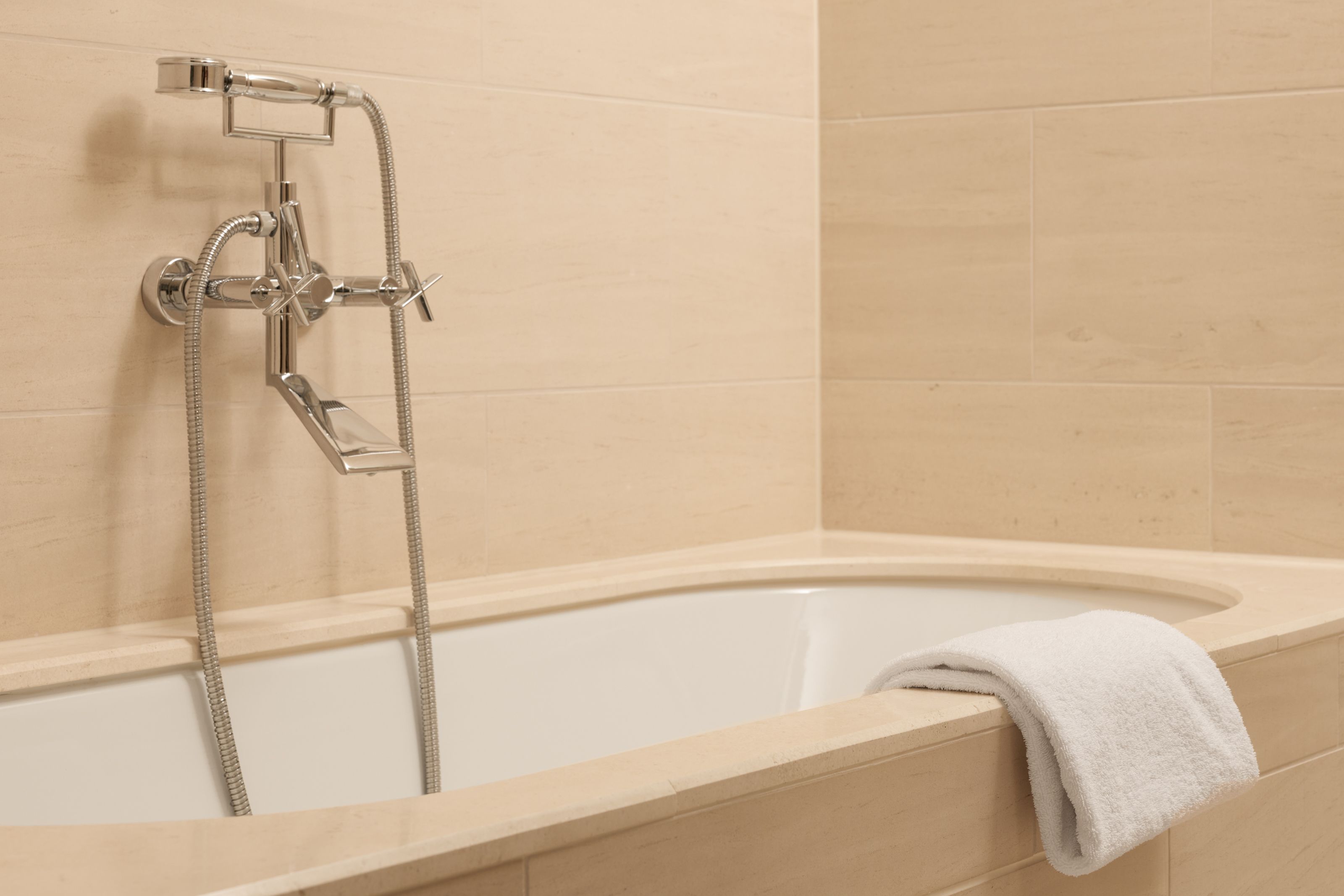 Freistehende Badewanne mit moderner verchromter Armatur und Handbrause, umgeben von beige gefliesten Wänden, mit einem weißen Handtuch am Wannenrand für entspannende Momente im Badezimmer.