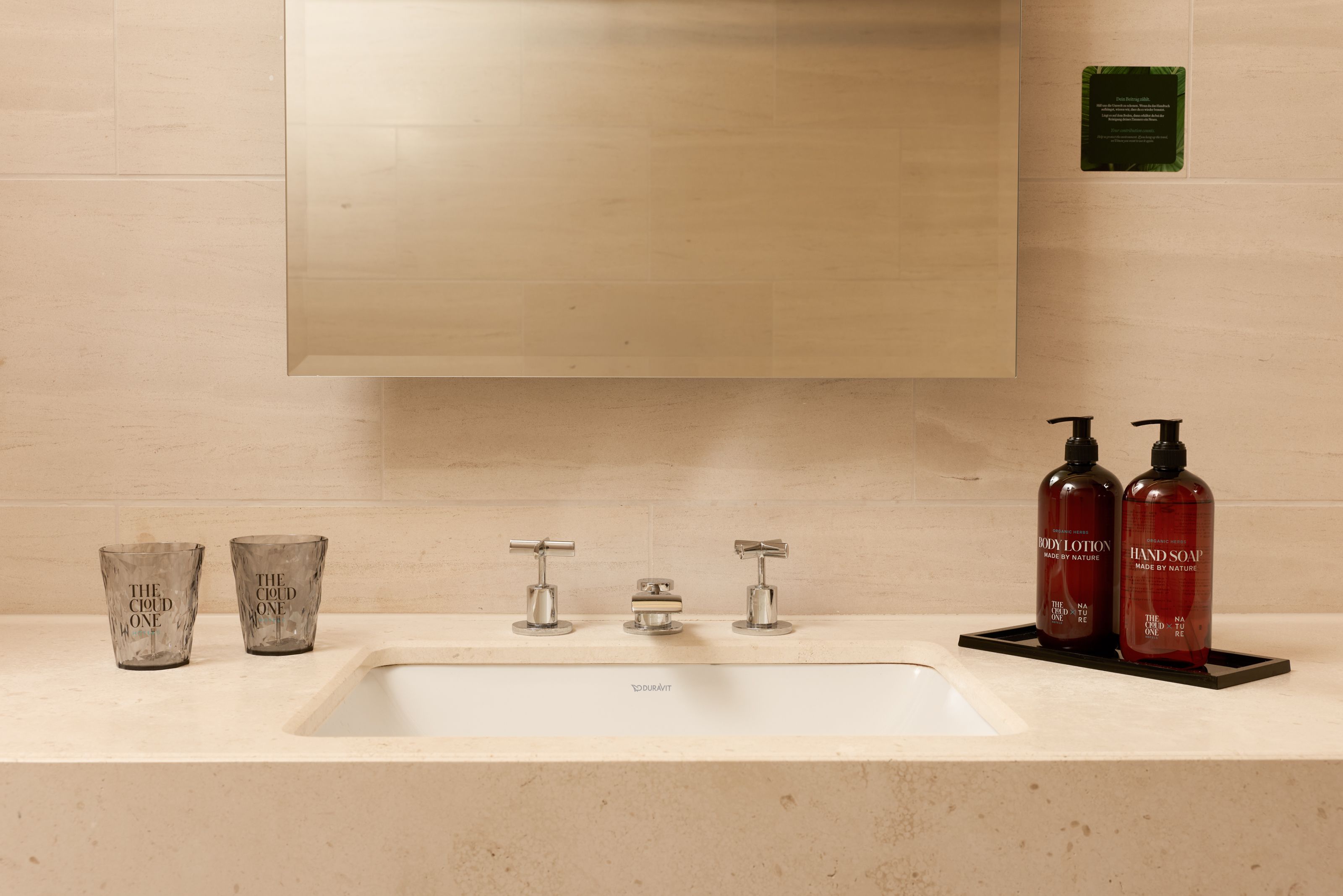 Moderner Badezimmerwaschbeckenbereich mit integriertem Spiegel, eleganten Wasserhähnen, zwei Seifenspendern und dekorativen Bechern auf hellem, sauberem Steinwaschtisch.