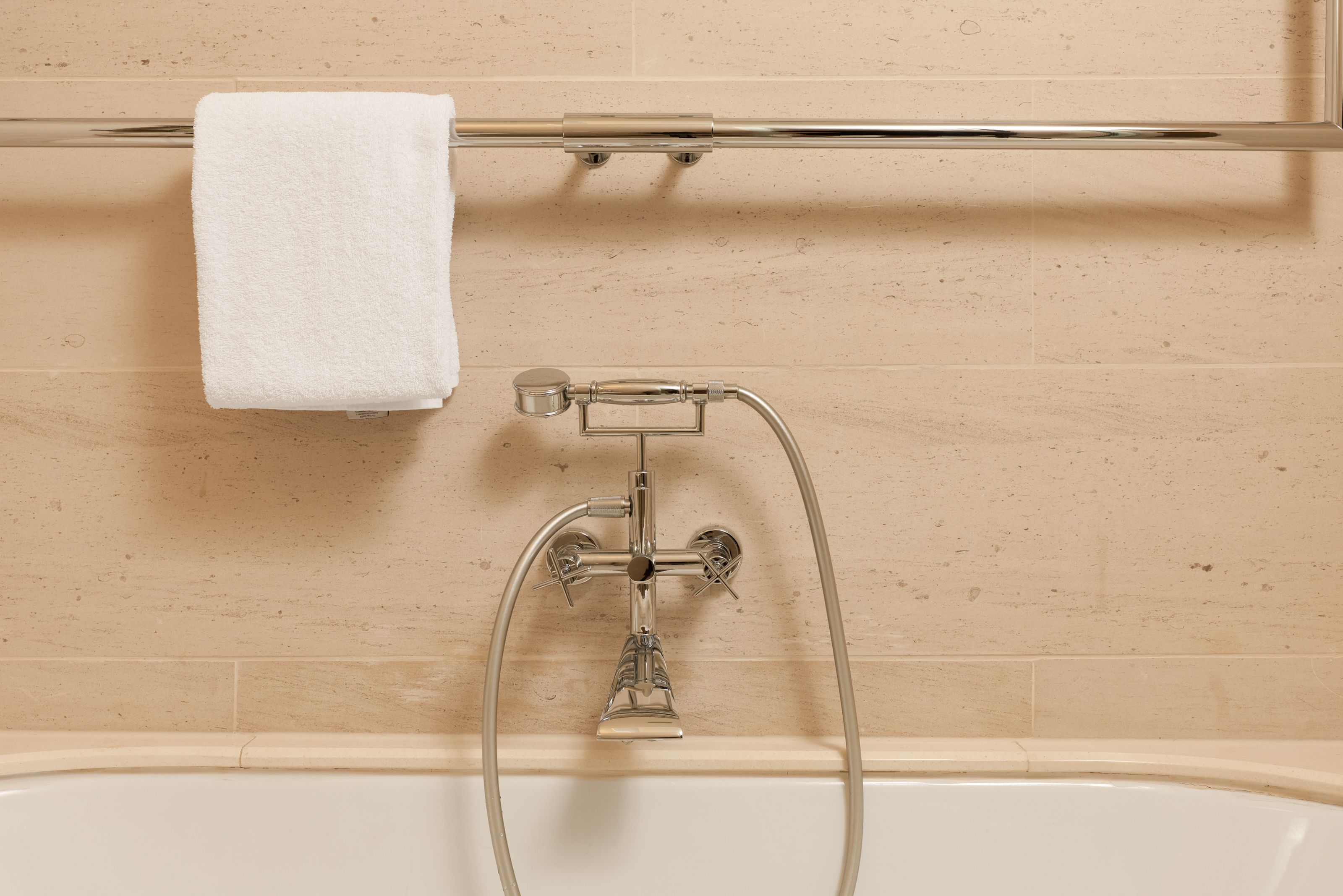 Moderne Badewannenarmatur in Chrom mit Handbrause an einer cremefarbenen Wand, darüber ein Handtuch an einem Handtuchhalter.