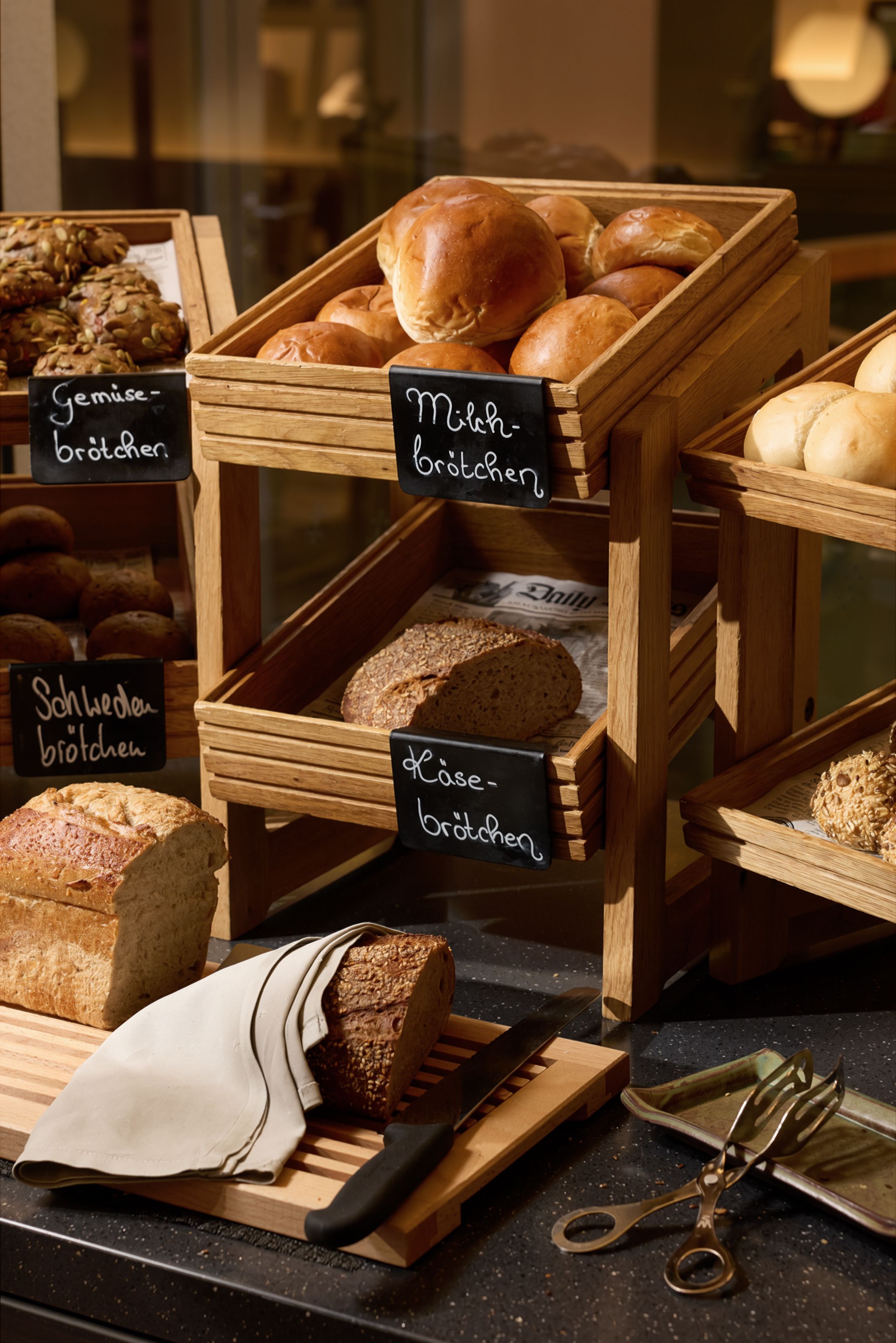 Verschiedene frische Brötchen und Brotsorten in Holzregalen, darunter Milchbrötchen, Käsebrötchen und Gemüsebrötchen, in einem warm beleuchteten Buffetbereich. Ein Messer und eine Brotlaib mit einem Tuch sind im Vordergrund zu sehen.