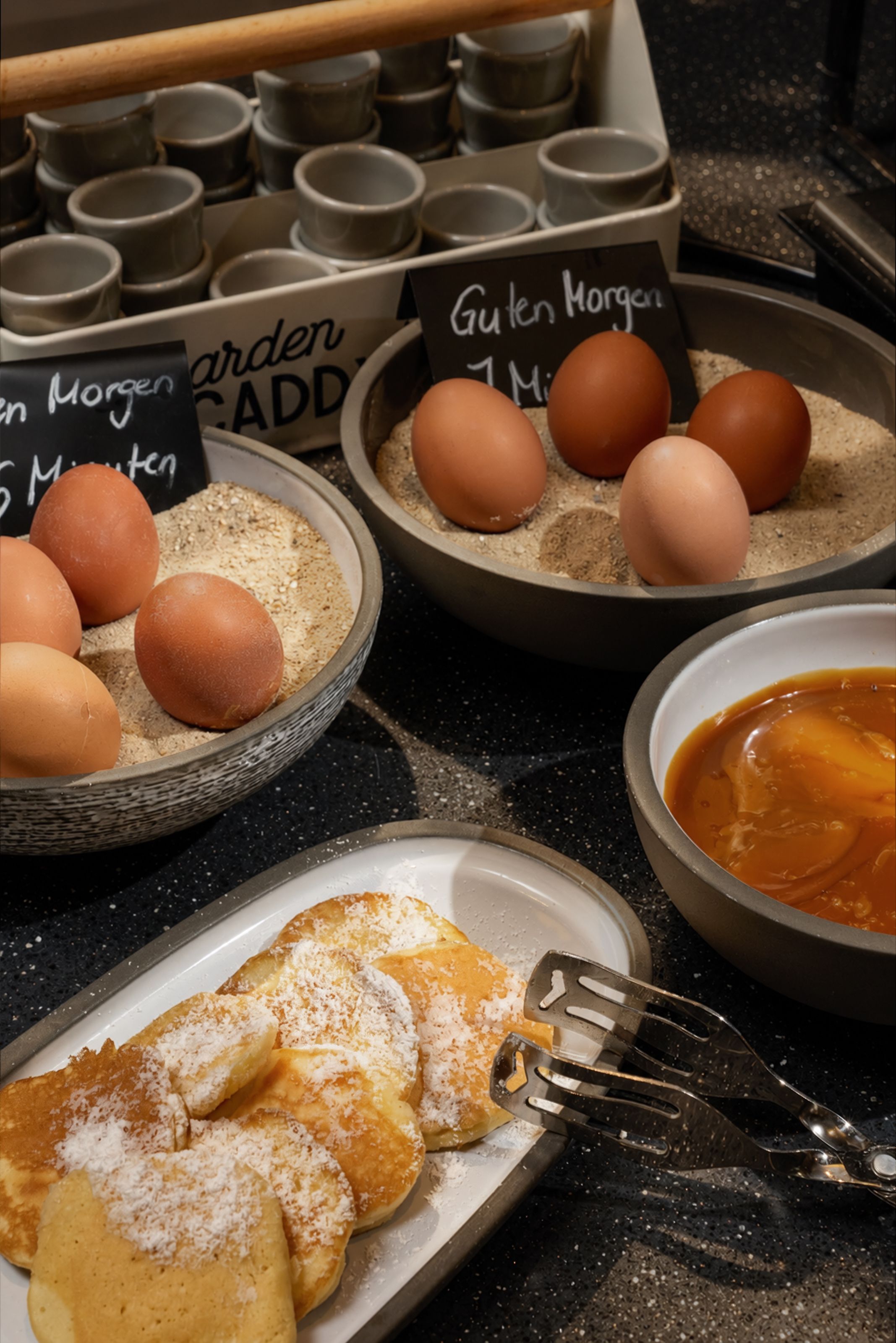 Frisch zubereitete Pfannkuchen mit Puderzucker, daneben Schalen mit Eiern in Sand, kennzeichnet als „Guten Morgen 1 Minute“ und kleine Tassen im Hintergrund, typisch für ein Frühstücksbuffet in gemütlicher Atmosphäre.