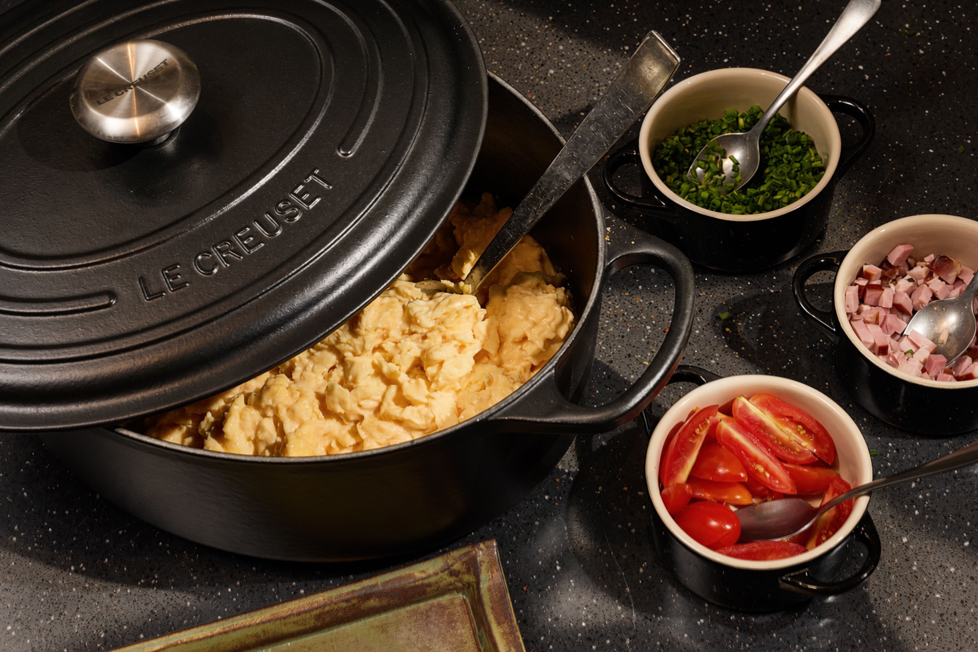 Schwarzer gusseiserner Topf mit cremigem Kartoffelpüree, daneben Schälchen mit gehackten Tomaten, Speckwürfeln und frischen Kräutern als Beilagen.