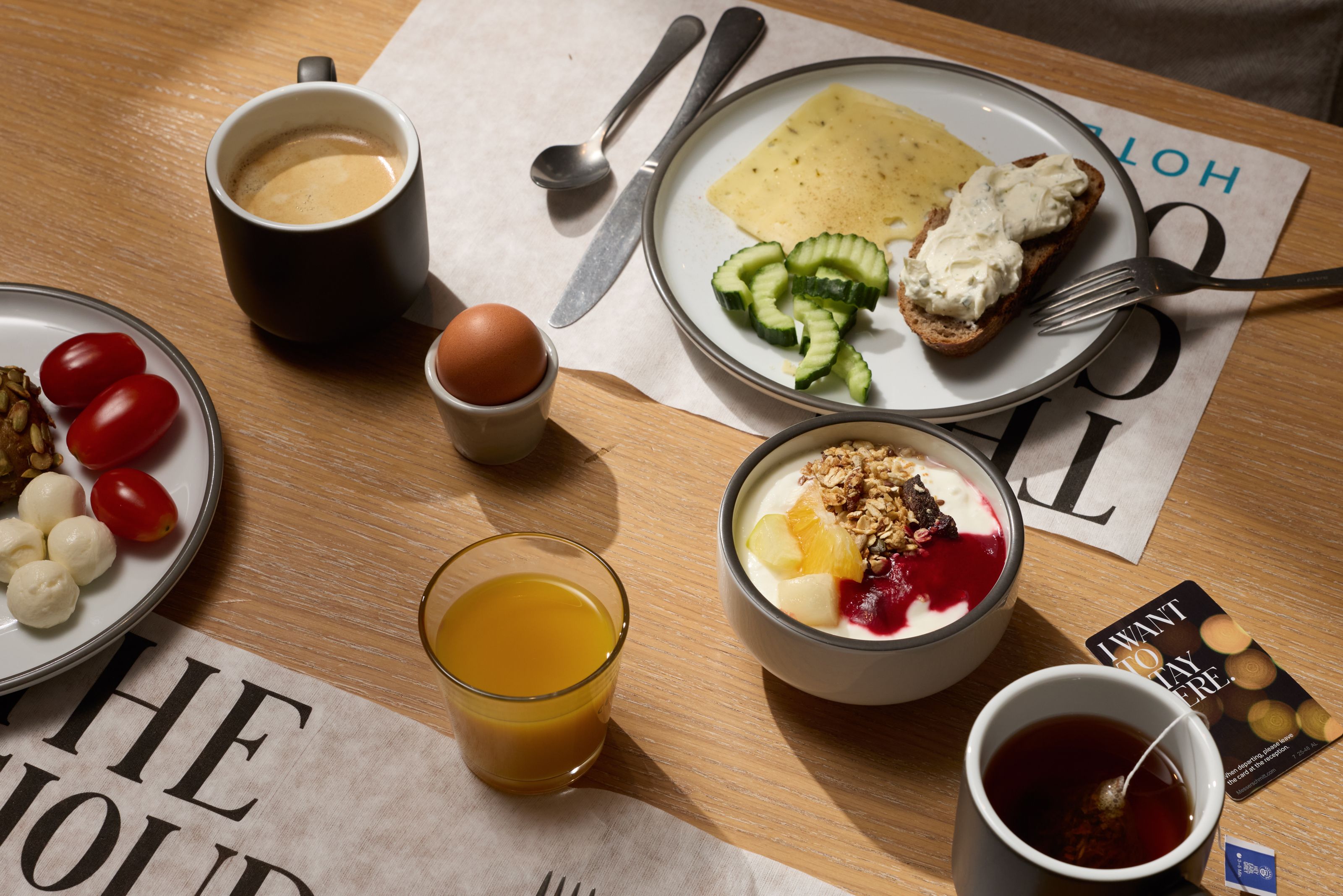 Frühstückstisch mit Kaffee, Tee, Orangensaft, gekochtem Ei, Joghurt mit Müsli und Beeren, Scheiben von Gurke, Avocado-Brot, Käse sowie Tomaten und Mozzarellakugeln.