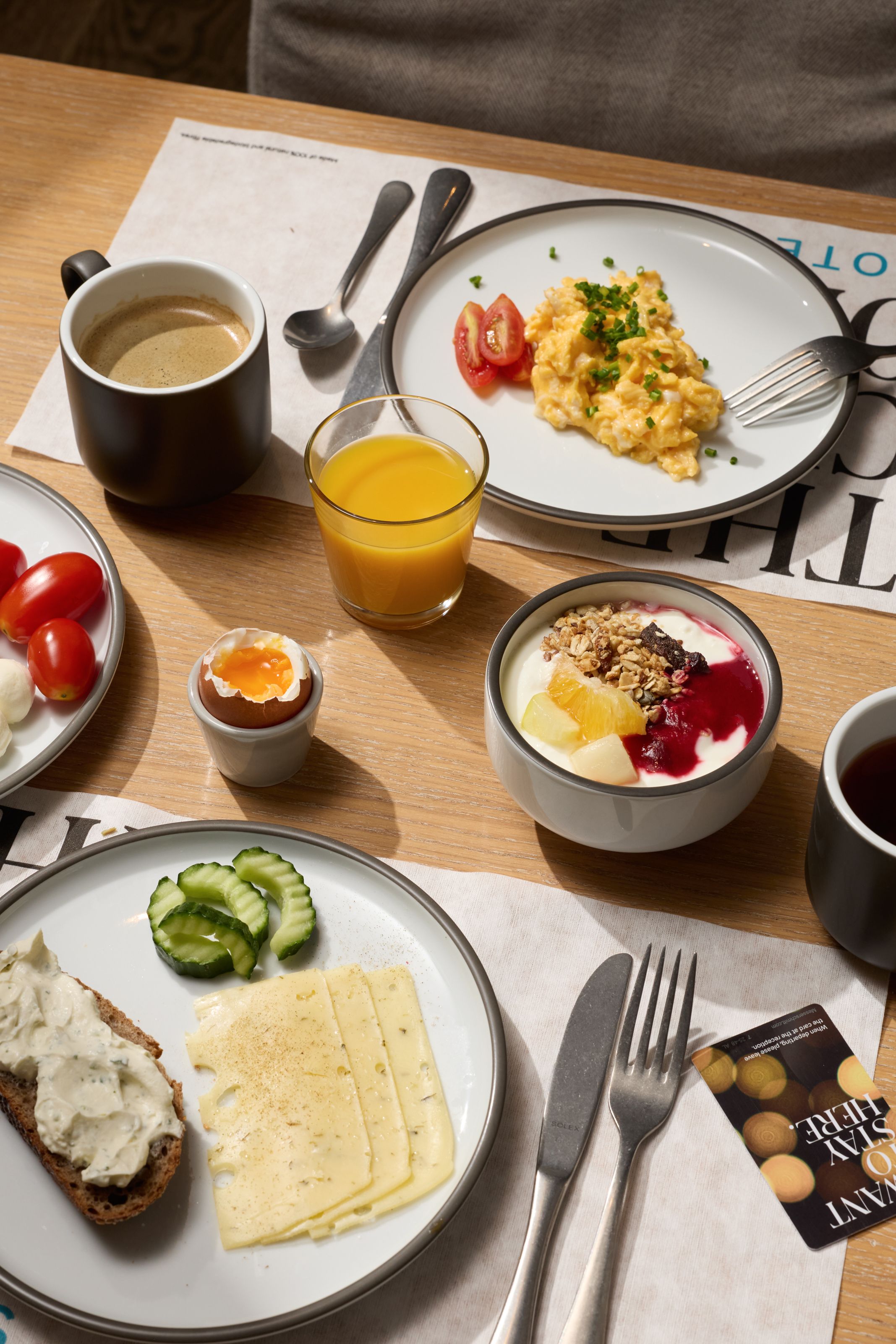 Frühstückstisch mit Rührei, Käse, Gurkenscheiben, Joghurt mit Früchten und Müsli, Kaffeetassen, Orangensaft, Marmelade und frischen Tomaten, alles ansprechend angerichtet auf hellem Holztisch.