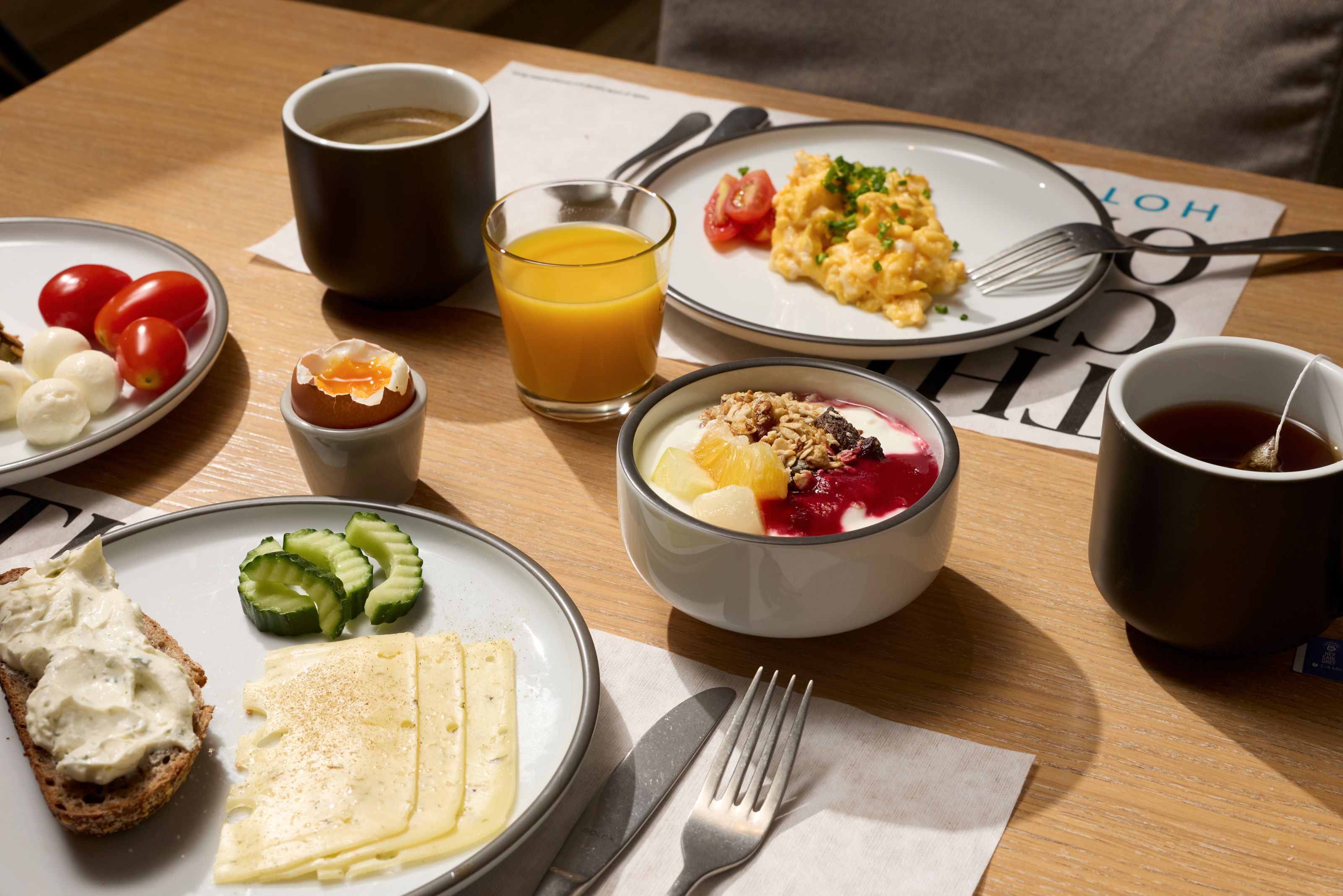 Frühstückstisch mit Rührei, Brot mit Frischkäse, Käse, Gurkenscheiben, Tomaten, gekochtem Ei, Joghurt mit Früchten und Müsli, Orangensaft, Kaffee und Tee, in gemütlicher Atmosphäre.
