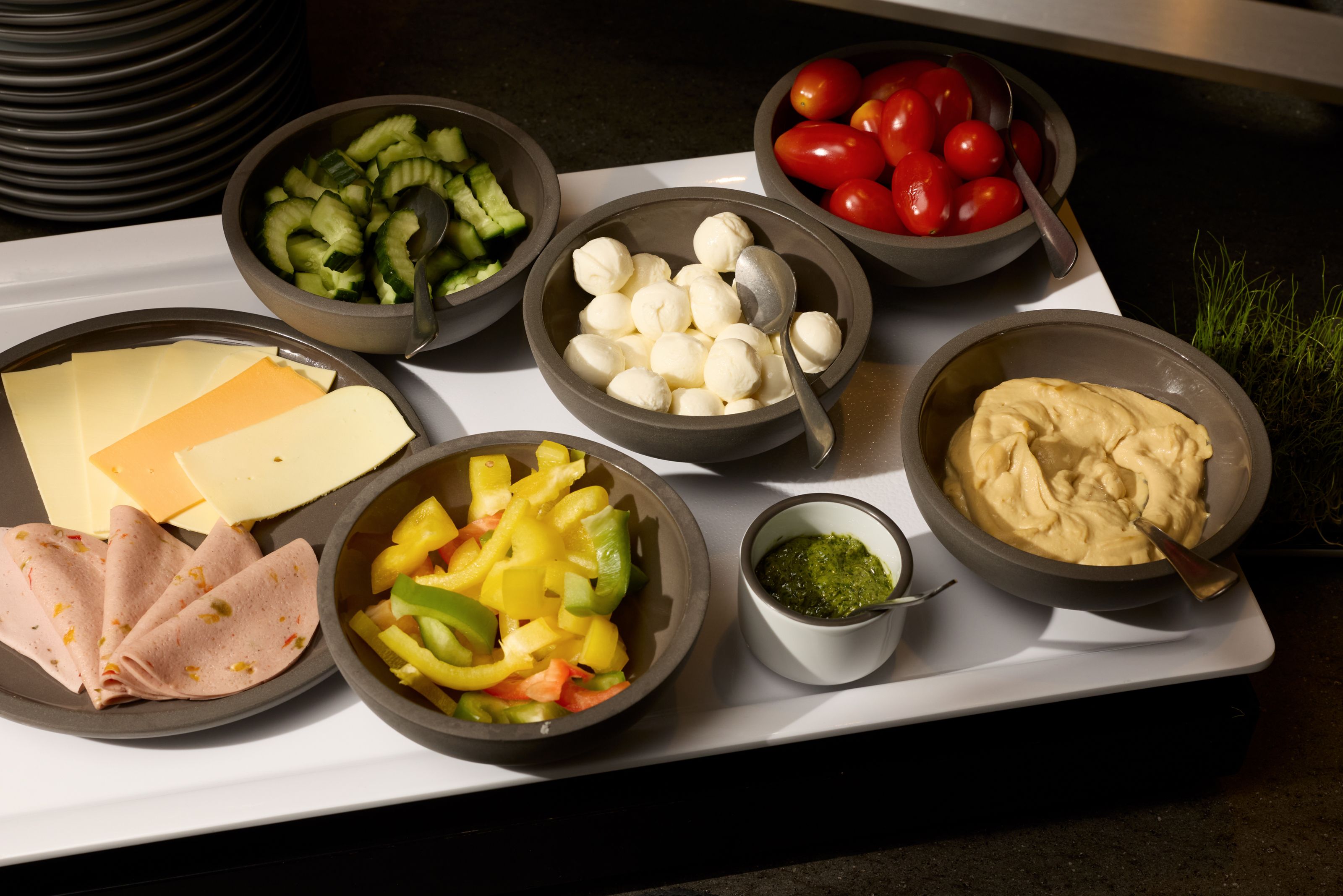 Verschiedene frische Zutaten wie Tomaten, Gurken, Mozzarella, Aufschnitt, Käse, bunte Paprika, Hummus und grünes Pesto, ansprechend in Schalen angerichtet, ideal für ein gesundes Frühstück oder Snack-Buffet.