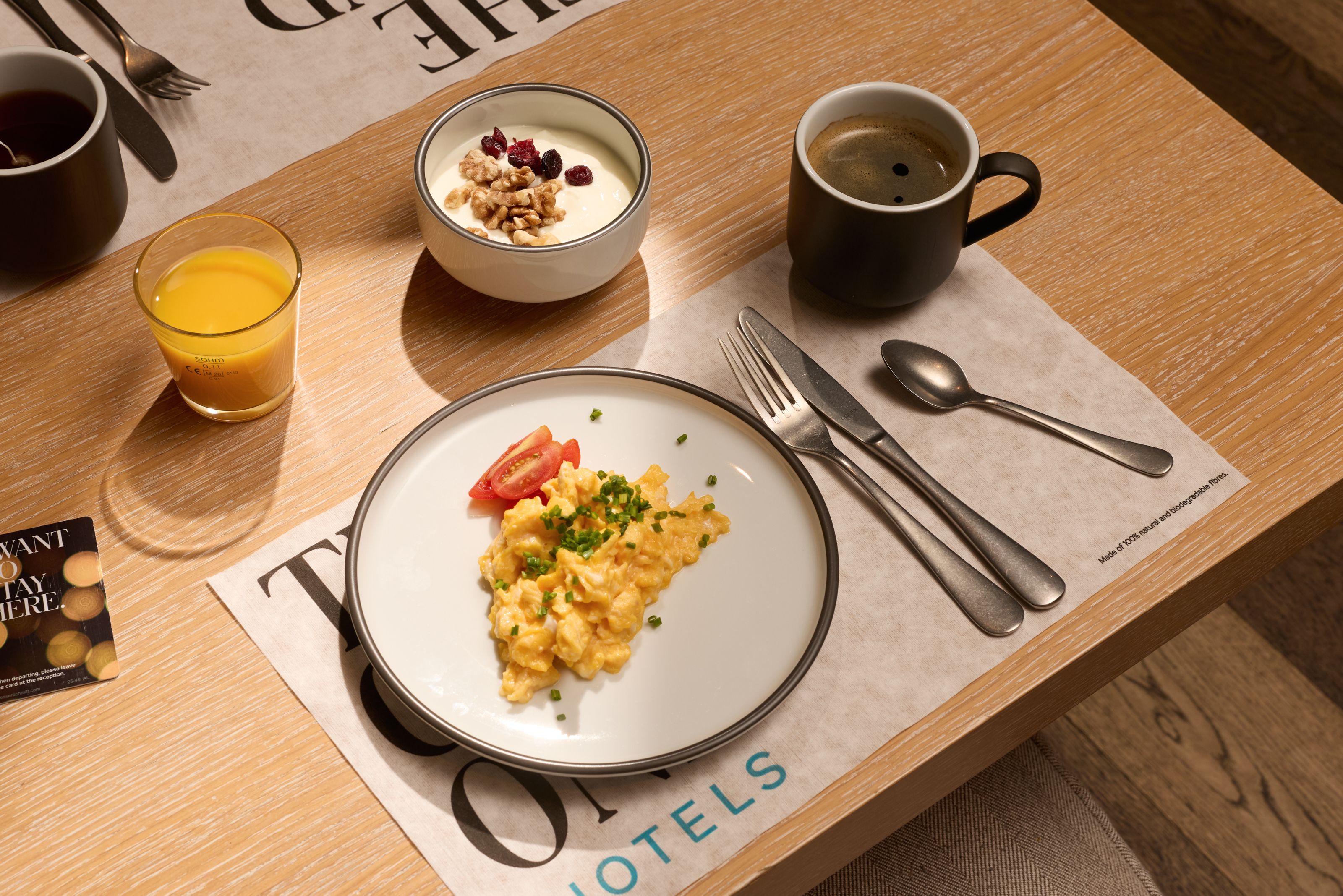 Ein gedeckter Frühstückstisch mit einem Teller Rührei, einem Glas Orangensaft, einer Schale Joghurt mit Nüssen und Cranberries sowie einer Tasse Kaffee, angerichtet auf einer hellen Holzoberfläche.