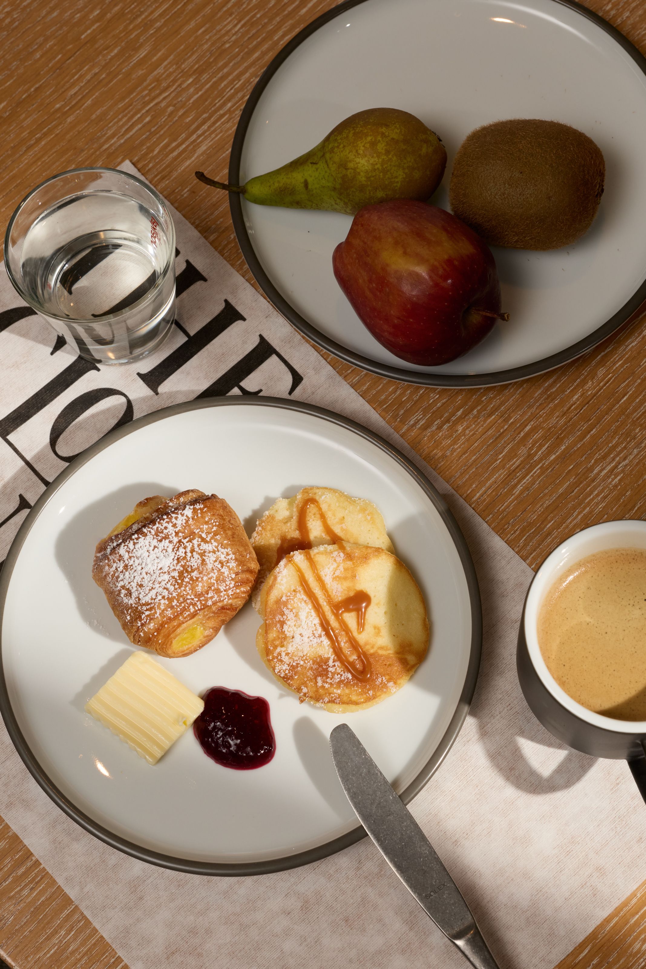 Frühstücksteller mit Pfannkuchen, Marmelade, Butter und einem Stück Gebäck, daneben eine Obstteller mit Birne, Apfel und Kiwi sowie eine Tasse Kaffee und ein Glas Wasser auf Holzoberfläche.