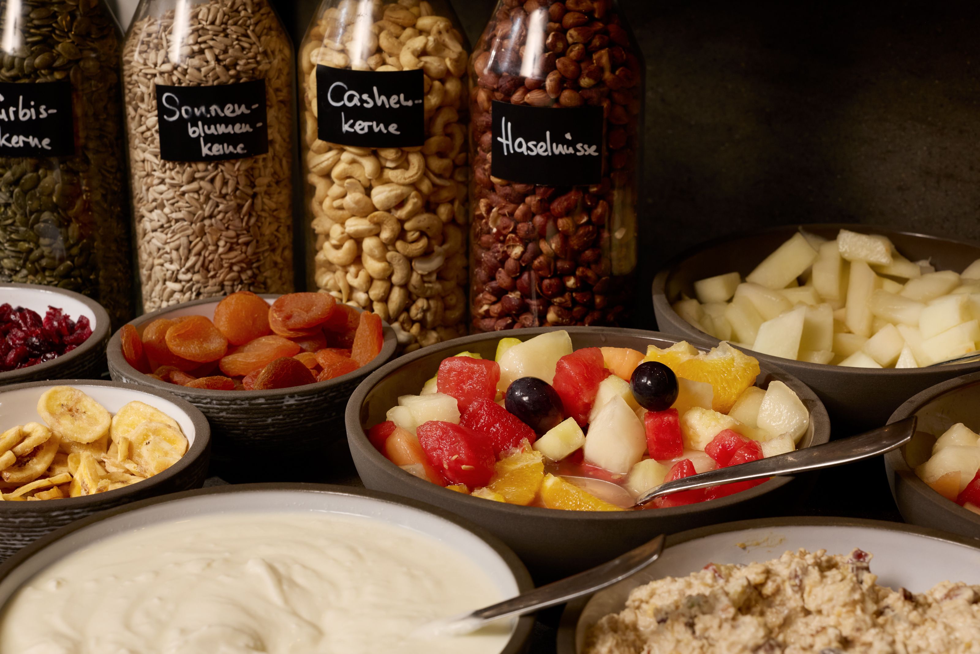 Verschiedene Schalen mit frischem Obstsalat, getrockneten Früchten, Joghurts und Nüssen, daneben Gläser mit Kürbiskernen, Sonnenblumenkernen, Cashewkernen und Haselnüssen für ein gesundes Frühstücksbuffet.