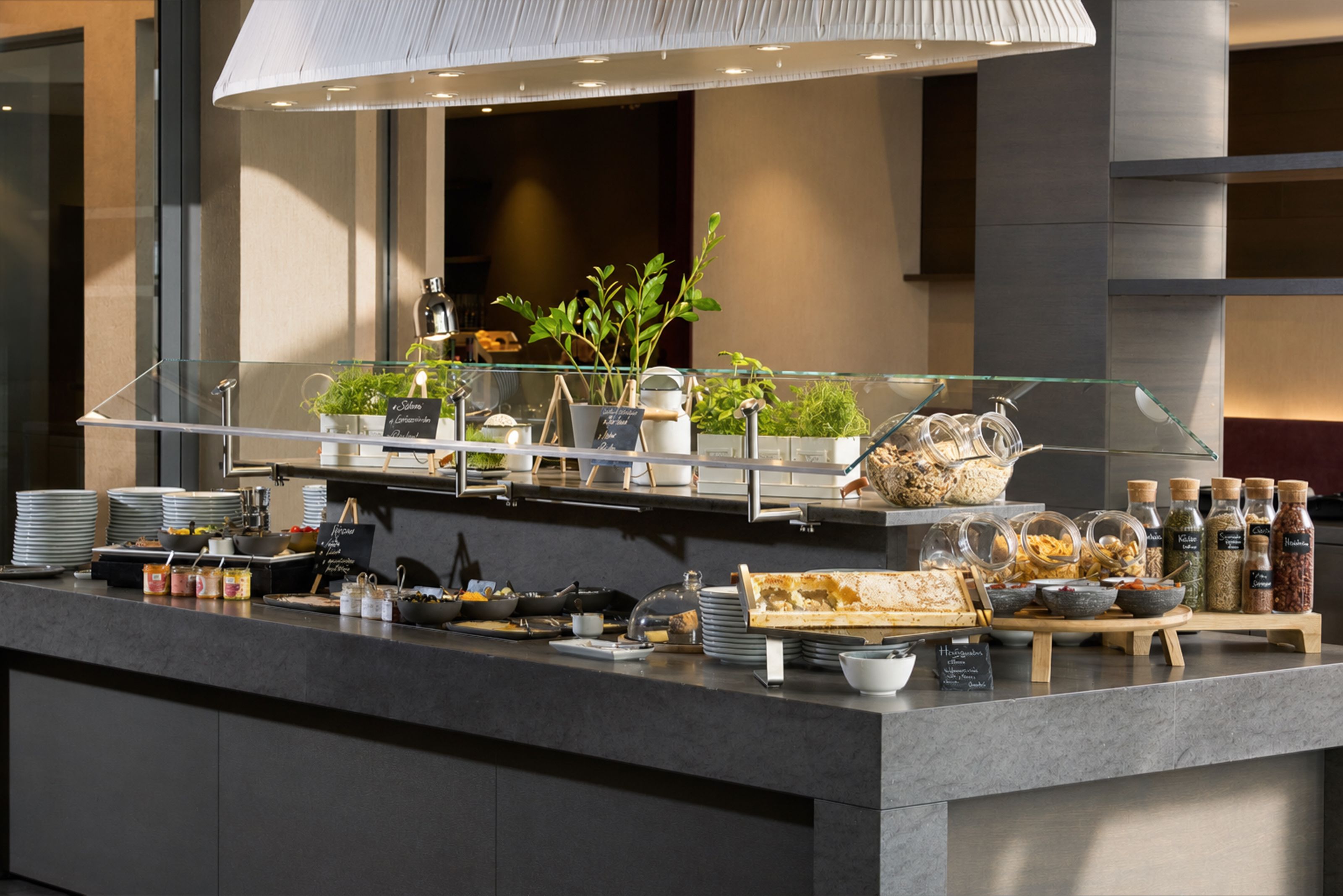 Buffetstation mit frischen Kräutern, verschiedenen Müslisorten, Brot und Frühstückszutaten, arrangiert auf modernem dunklem Tresen in hellem, einladendem Ambiente.