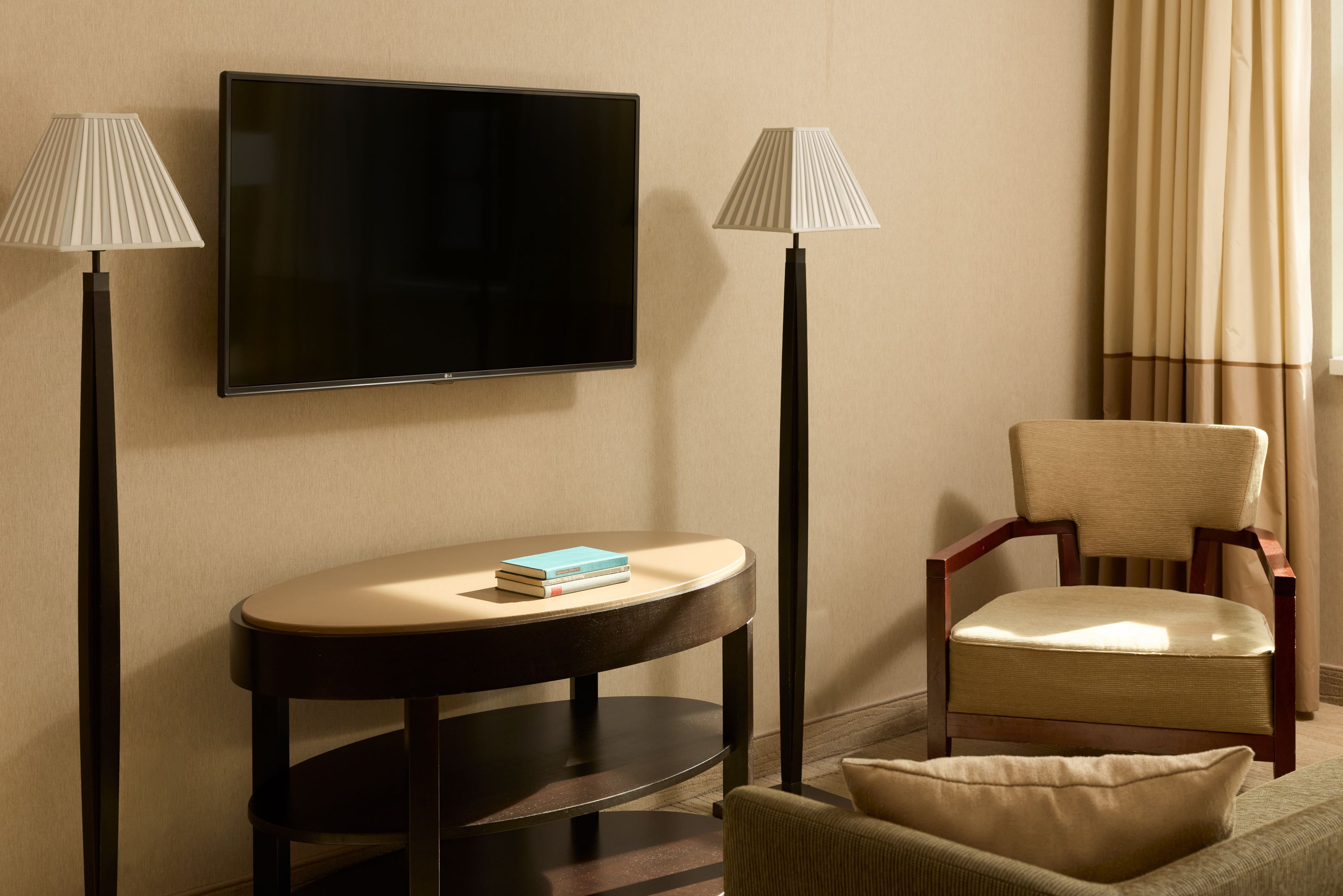 Helles, elegantes Hotelzimmer mit modernem Flachbildfernseher, zwei Stehlampen, einem ovalen Tisch mit Büchern und einem bequemen Sessel neben dem Fenster mit Vorhängen.