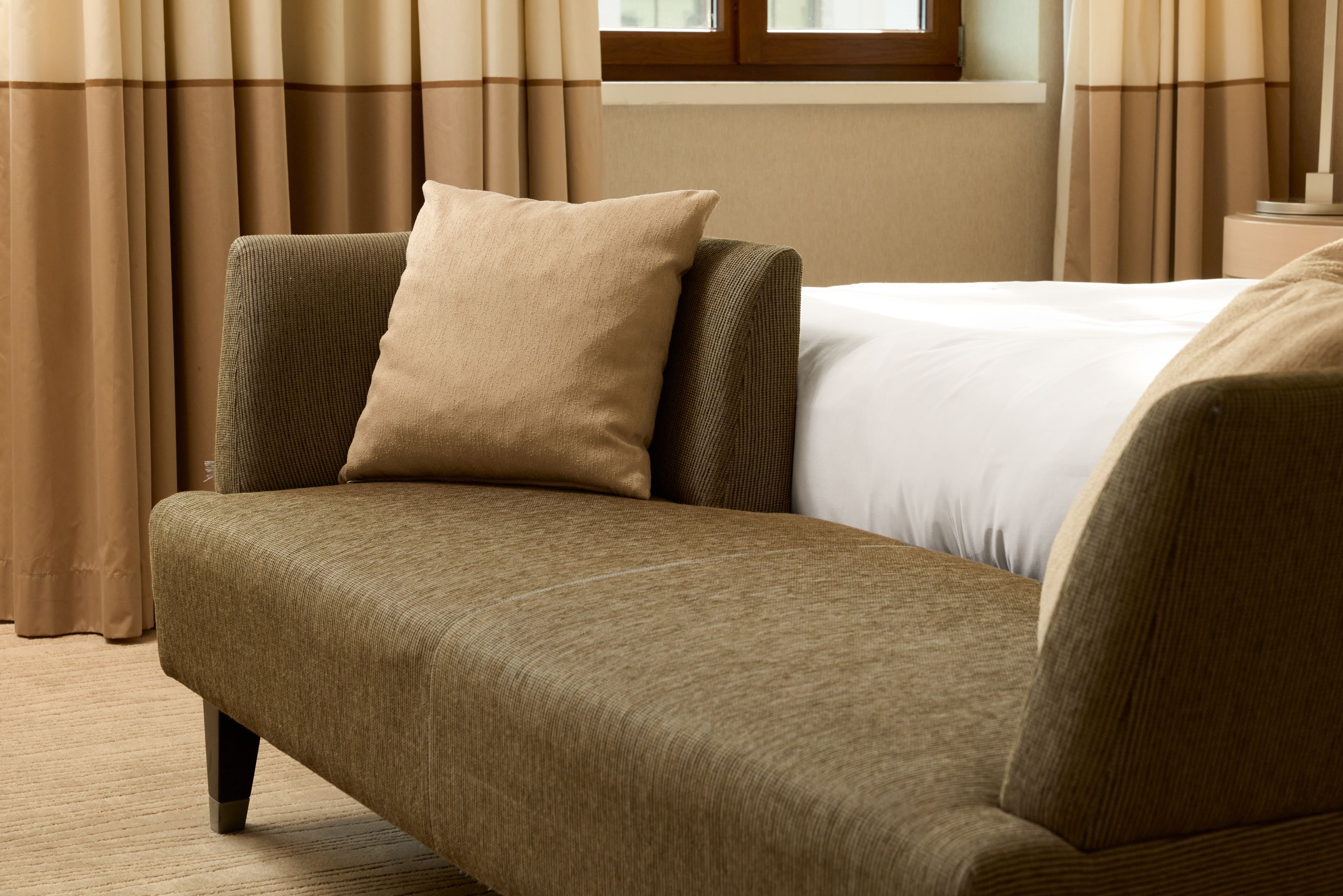 Beige-braunes Sofa mit Kissen in einem hellen Hotelzimmer neben einem Bett, vor einem Fenster mit braunen Gardinen, vermittelt eine gemütliche und ruhige Atmosphäre.