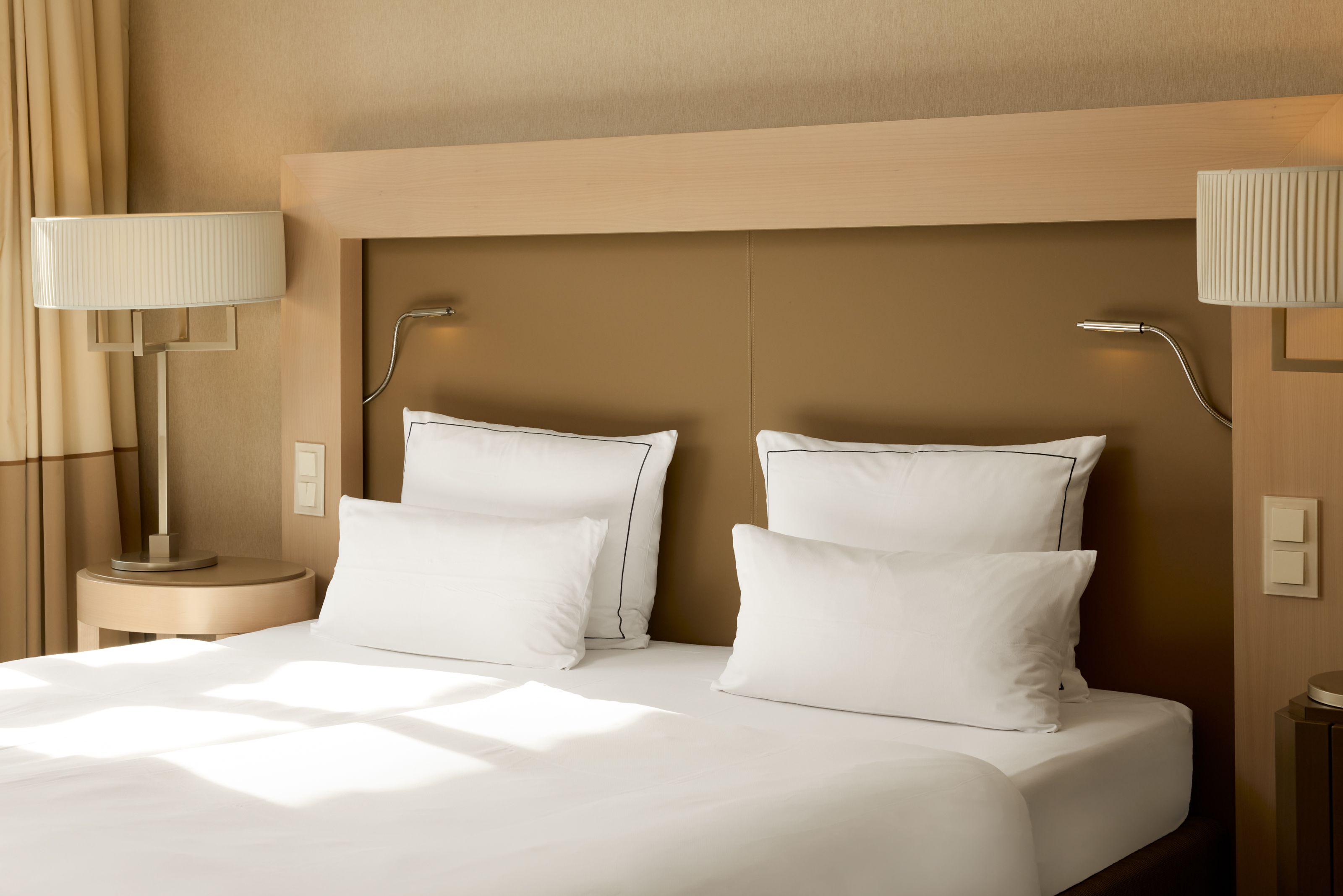 Hell eingerichtetes Hotelzimmer mit großem Doppelbett, weißen Kissen und Bettwäsche, modernem Kopfteil und stimmungsvoller Beleuchtung durch zwei Nachttischlampen auf beiden Seiten.