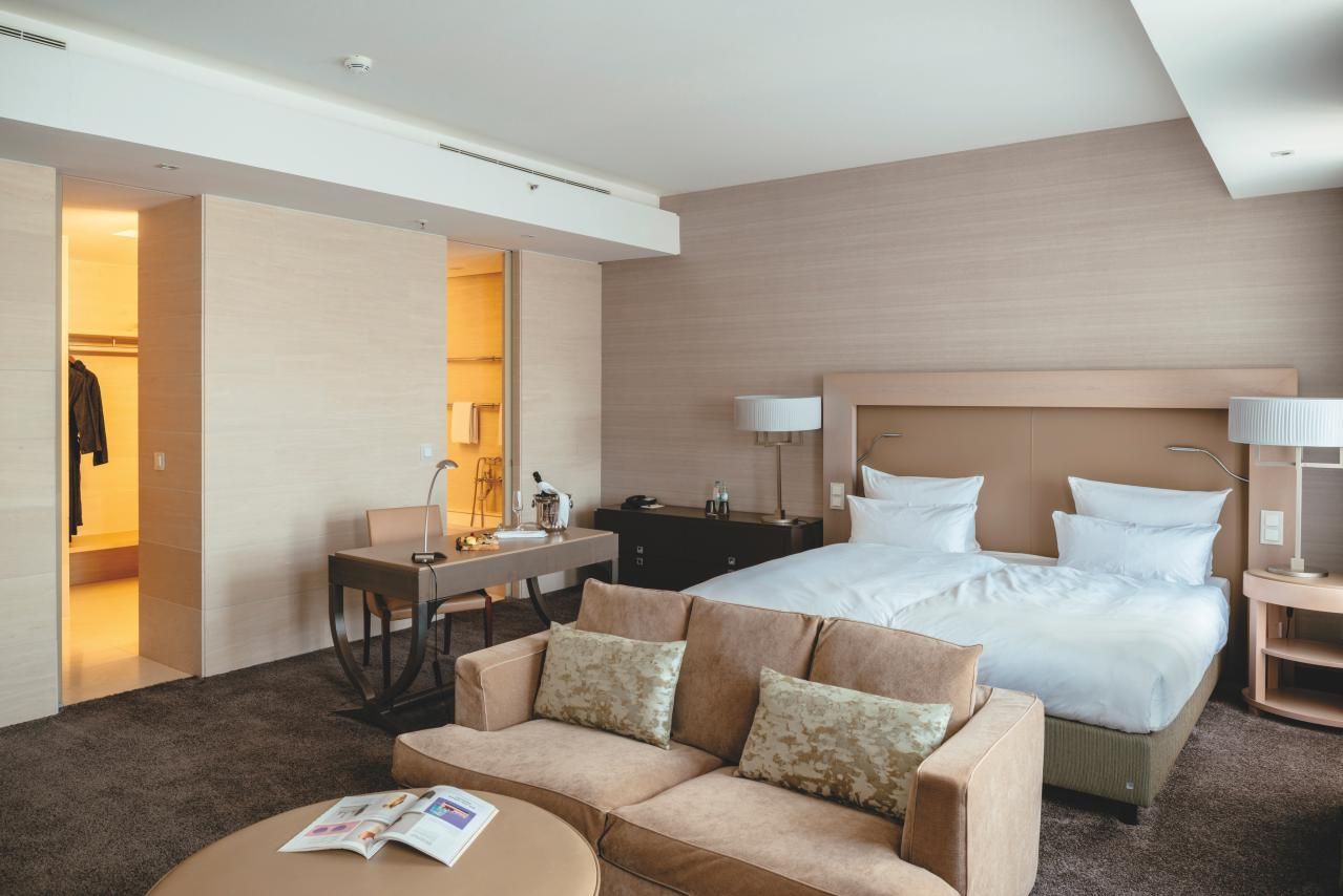 Modern eingerichtetes Hotelzimmer mit großem Doppelbett, gemütlichem Sofa, Arbeitsplatz und offenem Kleiderschrank, freundliche Beleuchtung und warme Farbtöne für entspannte Atmosphäre.