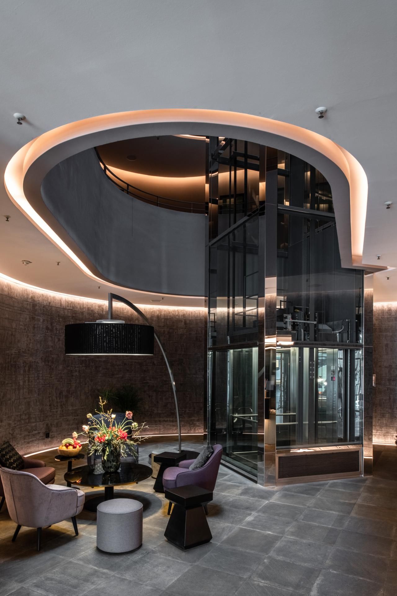 Moderner Hotel-Loungebereich mit Sitzmöbeln, Blumenarrangement und indirekter Beleuchtung, umgeben von einer dekorativen Deckenstruktur und einem gläsernen Aufzug, der Eleganz und komfortable Atmosphäre verbindet.