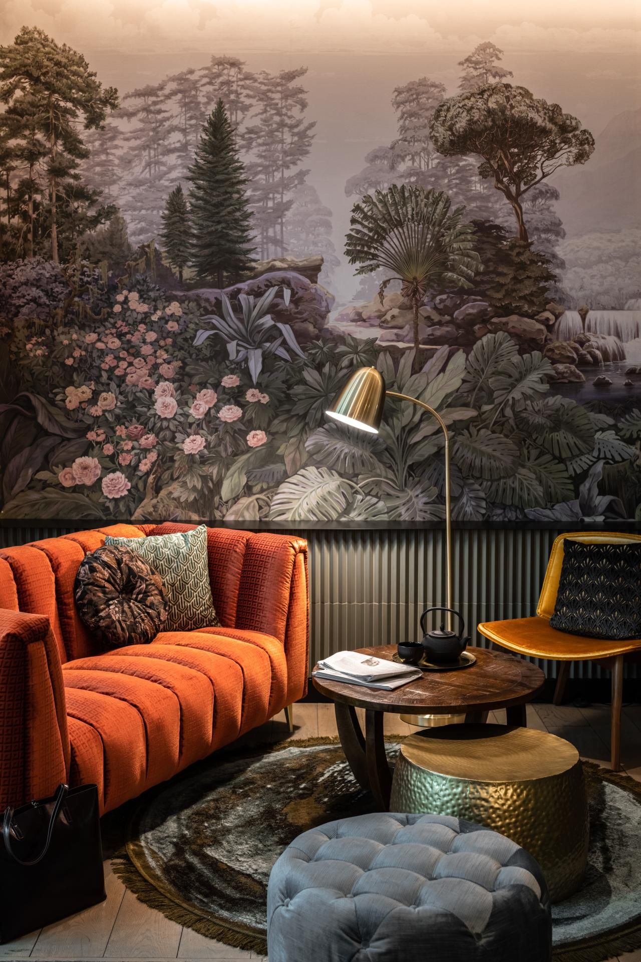 Gemütliche Lounge mit orangefarbenem Sofa, runder Kaffeetisch und Stehlampe vor einer Wand mit großem, detailliertem Naturkunstwerk, das Bäume und Pflanzen in beruhigenden Farben zeigt. Moderne Einrichtung mit warmem, einladendem Ambiente.