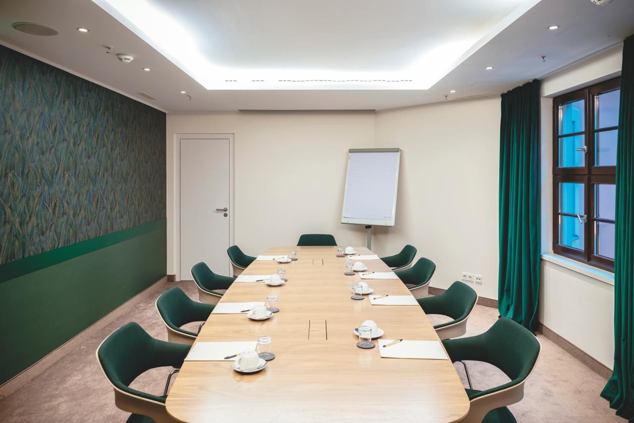 Konferenzraum mit langem Holztisch, umgeben von grünen Stühlen, mit Notizblöcken, Tassen und Wasser; helle, moderne Atmosphäre, Fenster mit grünem Vorhang und Flipchart an einer Wand.