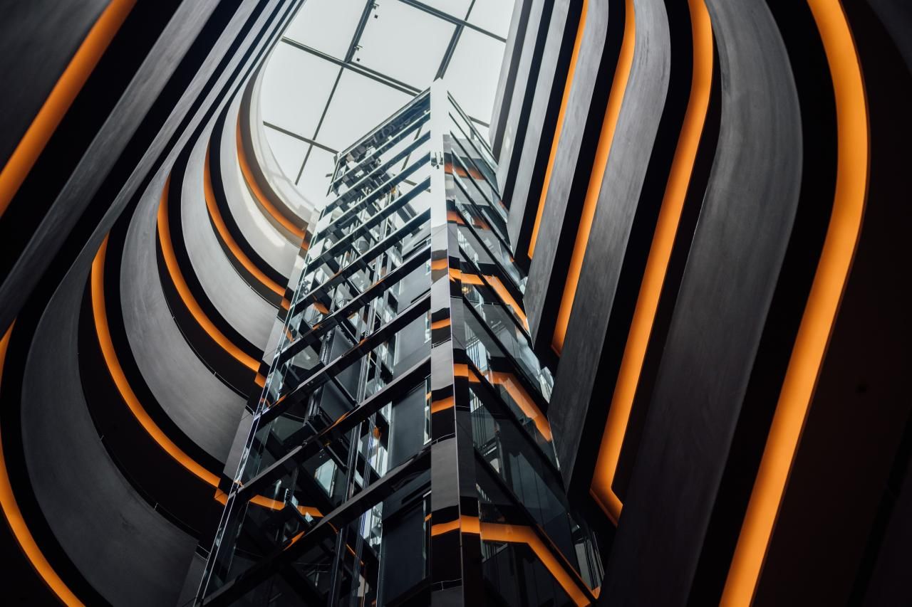 Blick nach oben in einem modernen Atrium mit einem gläsernen Aufzug in der Mitte und gebogenen Wänden, die mit warmen orangen Lichtlinien akzentuiert sind. Ein großes Oberlicht sorgt für Tageslicht.