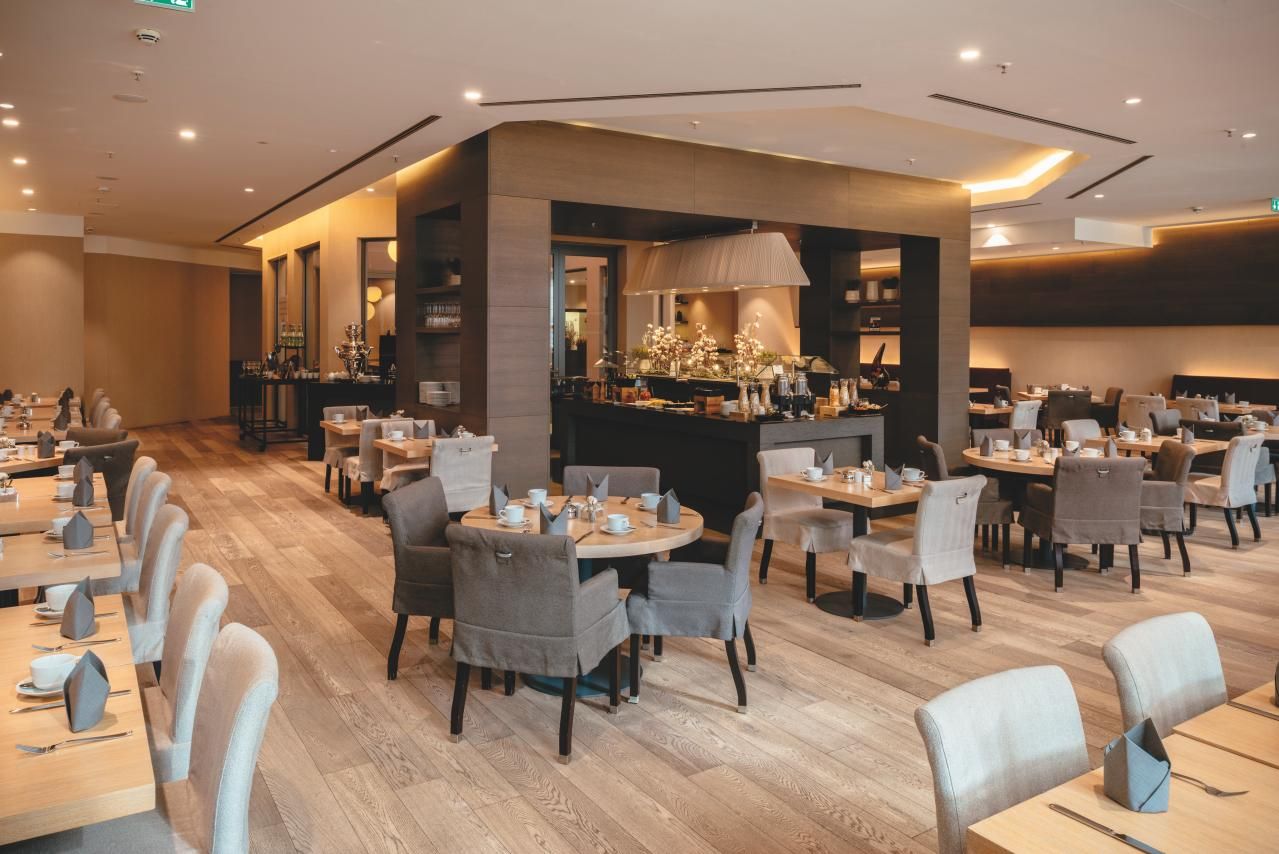Modernes Restaurant mit hellen Holzböden und gemütlichen Sesseln, großzügig angeordnete Tische, angenehme Beleuchtung, Buffetbereich in der Mitte, einladende Atmosphäre für entspanntes Essen und gesellige Runden.