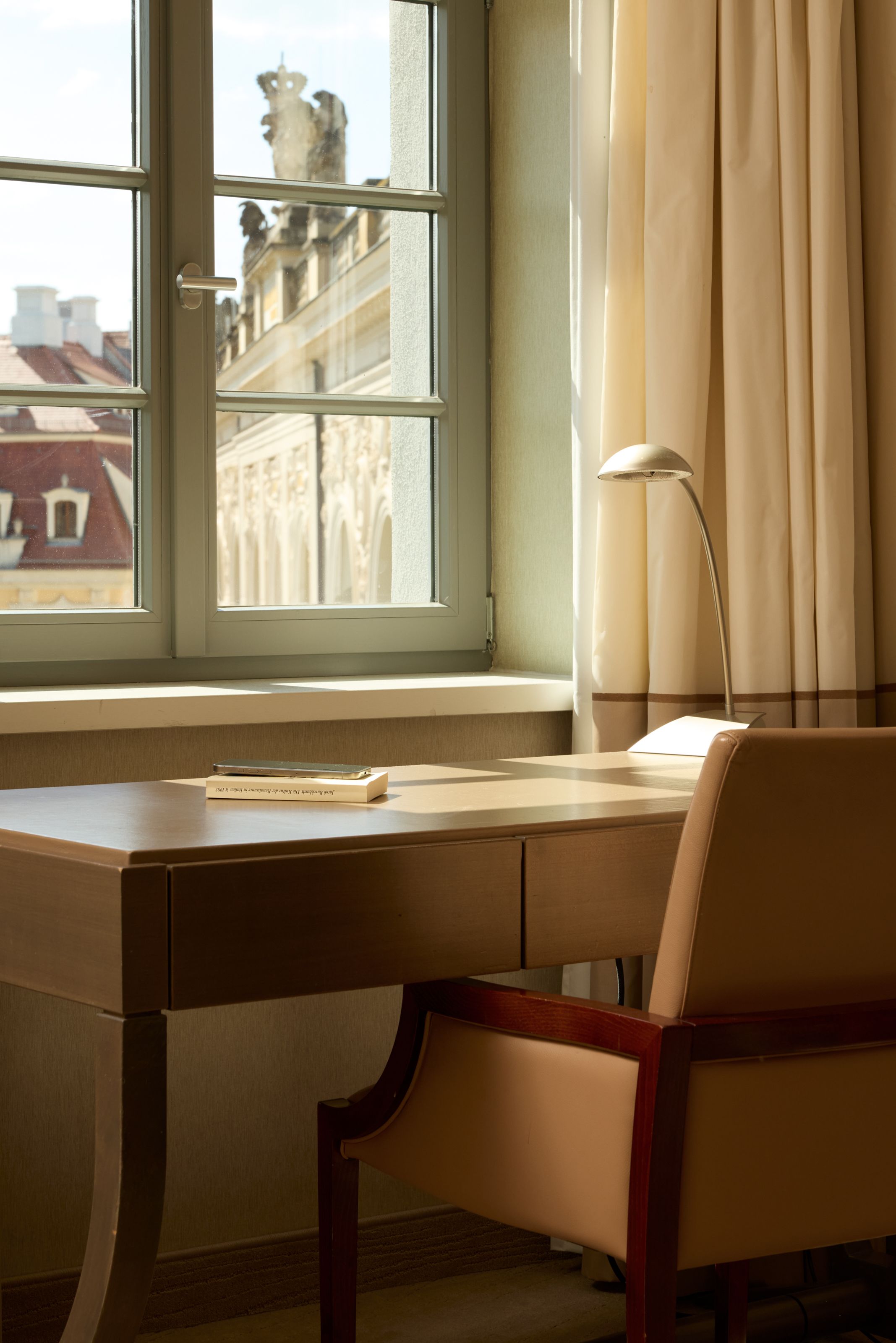 Heller Arbeitsplatz mit Schreibtisch, bequemer Ledersessel und moderner Lampe vor einem Fenster. Durch das Fenster Blick auf historische Gebäude mit verzierten Fassaden und Skulpturen. Ruhige, einladende Atmosphäre zum Arbeiten oder Lesen.