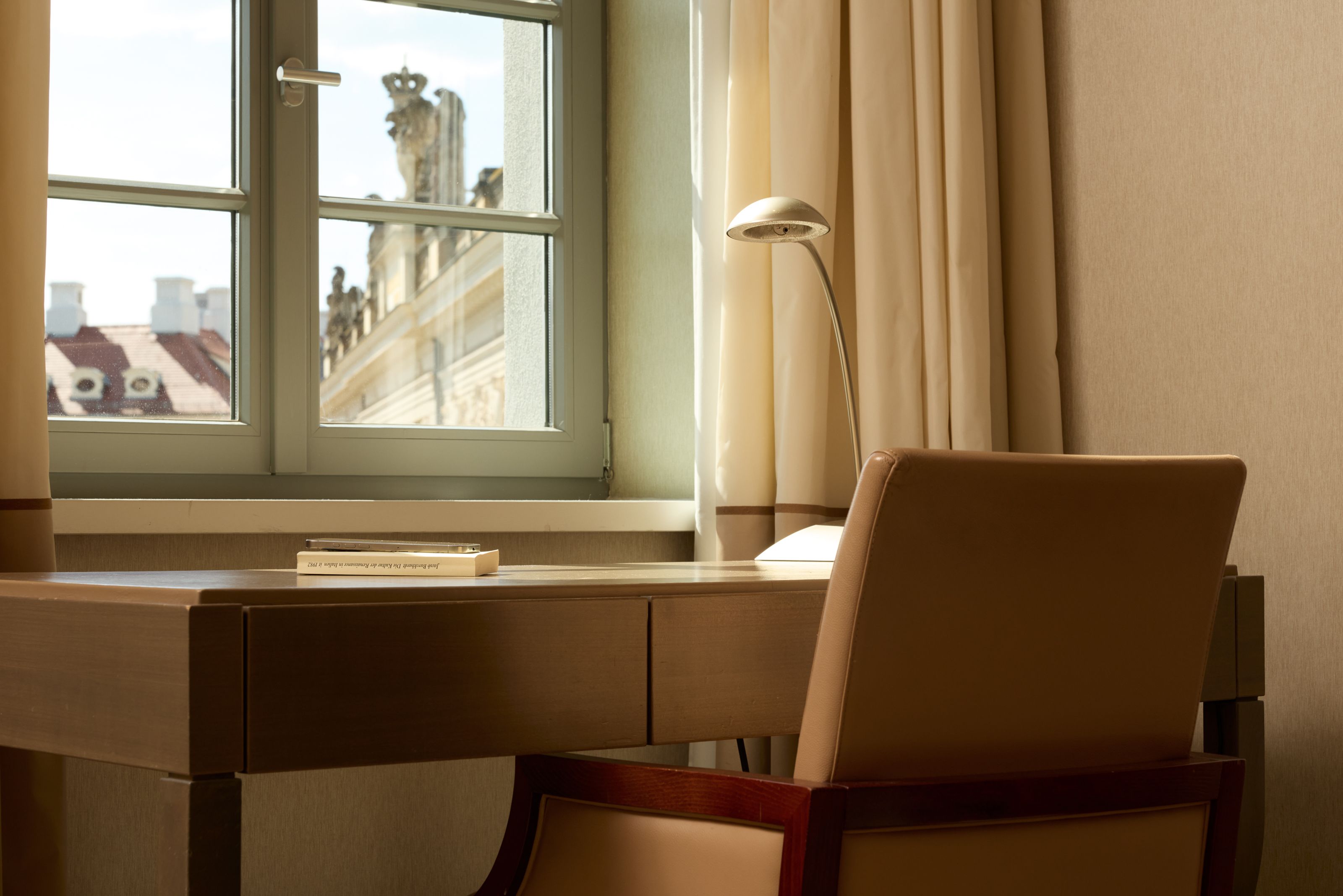 Heller Arbeitsplatz mit Holzschreibtisch und bequemen Ledersessel am Fenster, das eine historische Fassade und Sonnenschein zeigt. Ideal für ruhiges Arbeiten mit angenehmer Aussicht.