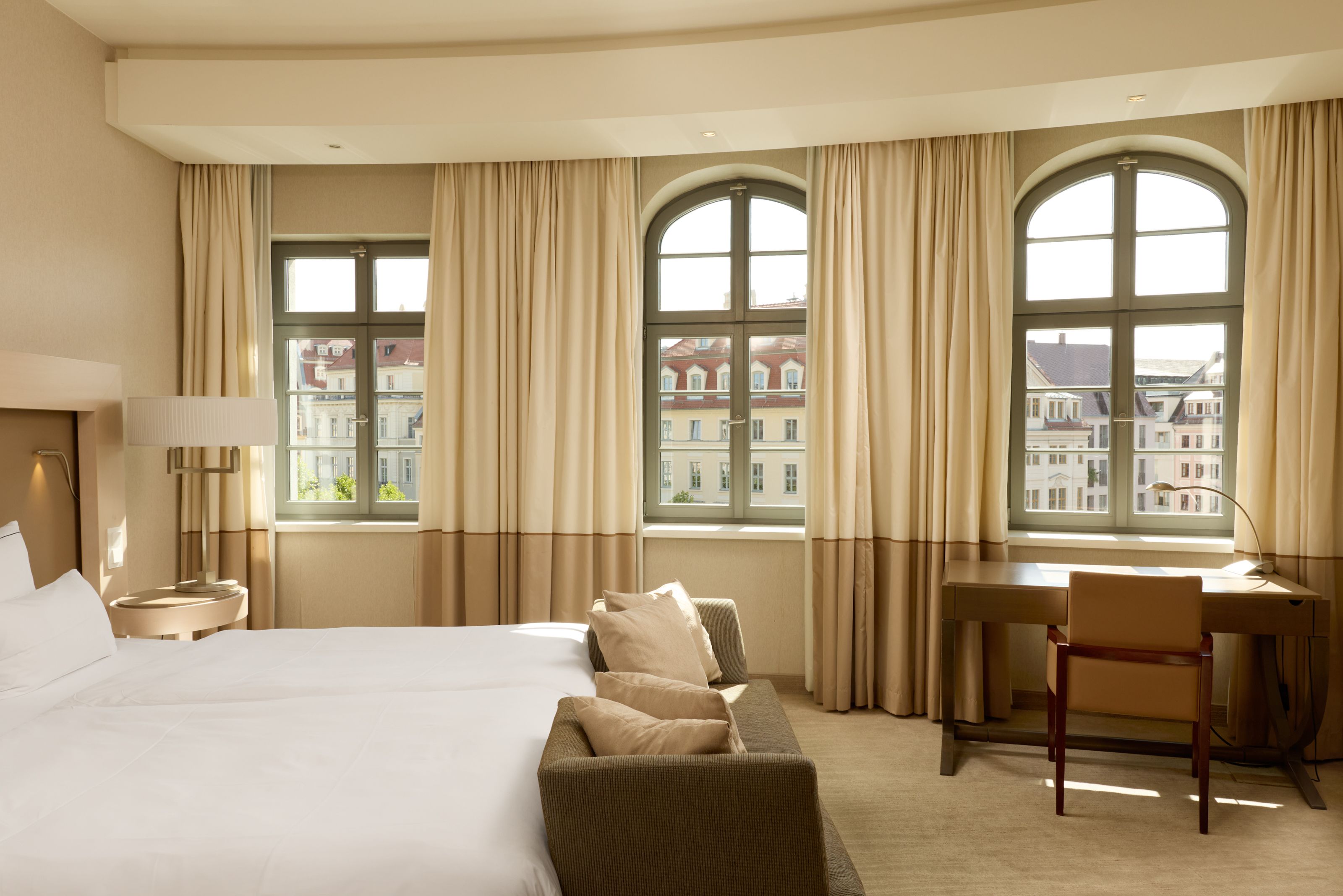 Helles Hotelzimmer mit großem Bett, Sofa, Schreibtisch und Stuhl. Drei große Fenster bieten Blick auf eine Altstadt mit historischen Gebäuden. Helle Vorhänge und warme Farben schaffen eine freundliche und einladende Atmosphäre.